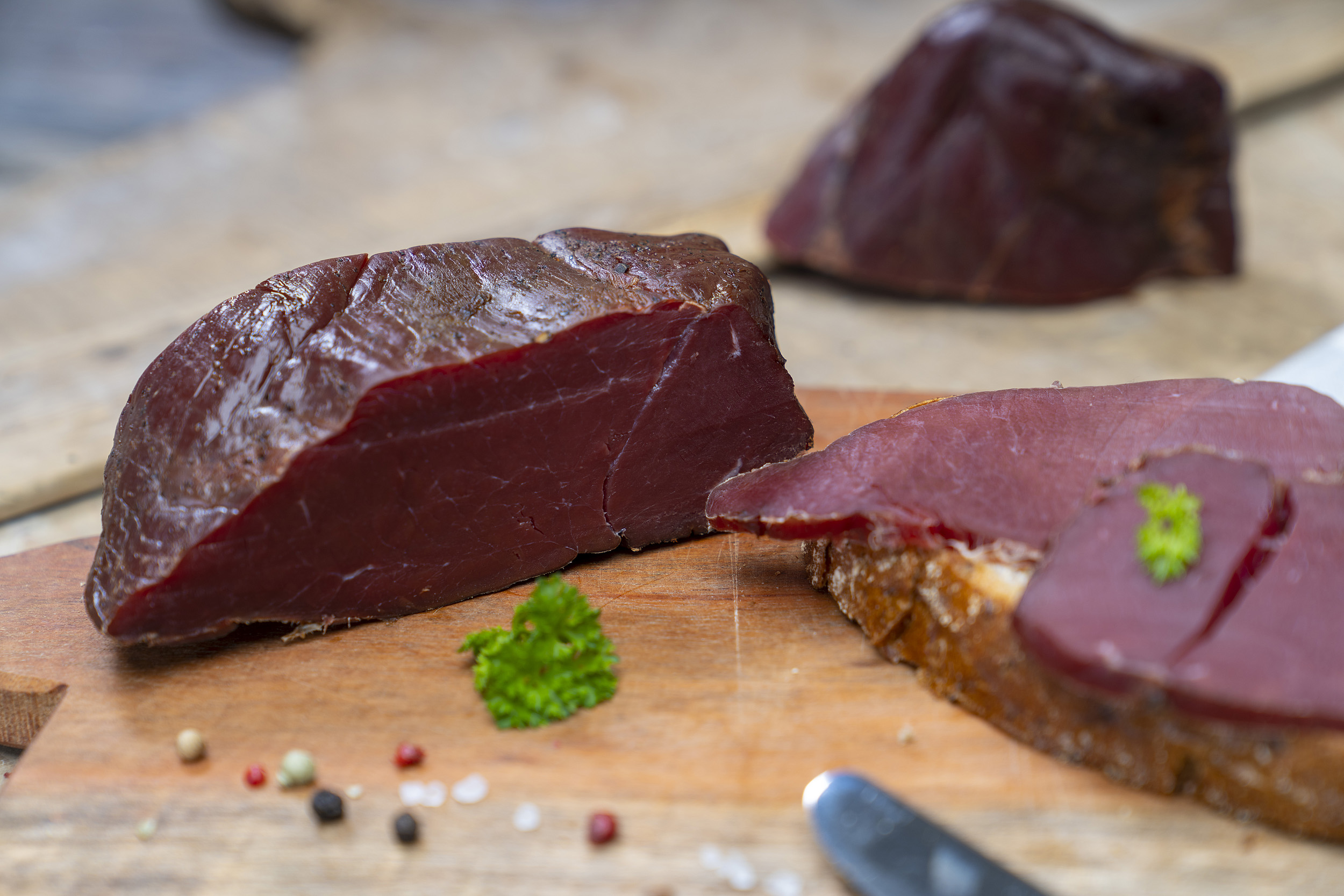 Fessels Wildschinken auf einem Brett. Daneben ein paar Scheiben und Gewürze. Hinten ein weiterer Schinken.