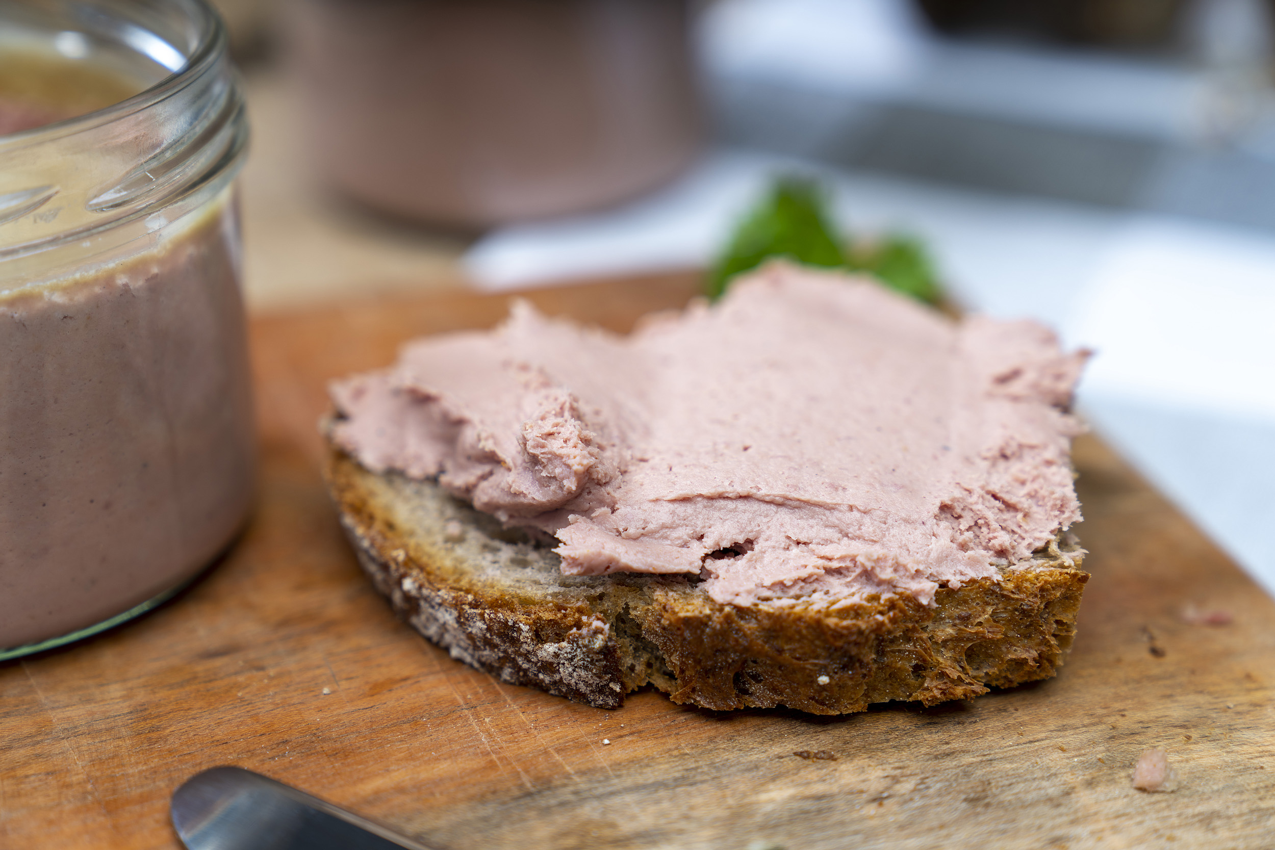 Nahaufnahme von einer geschmierten Schnitte mit Kalbfleischleberwurst. Daneben ein Glas mit der Leberwurst. Im Hintergrund Petersilie auf dem Brett.
