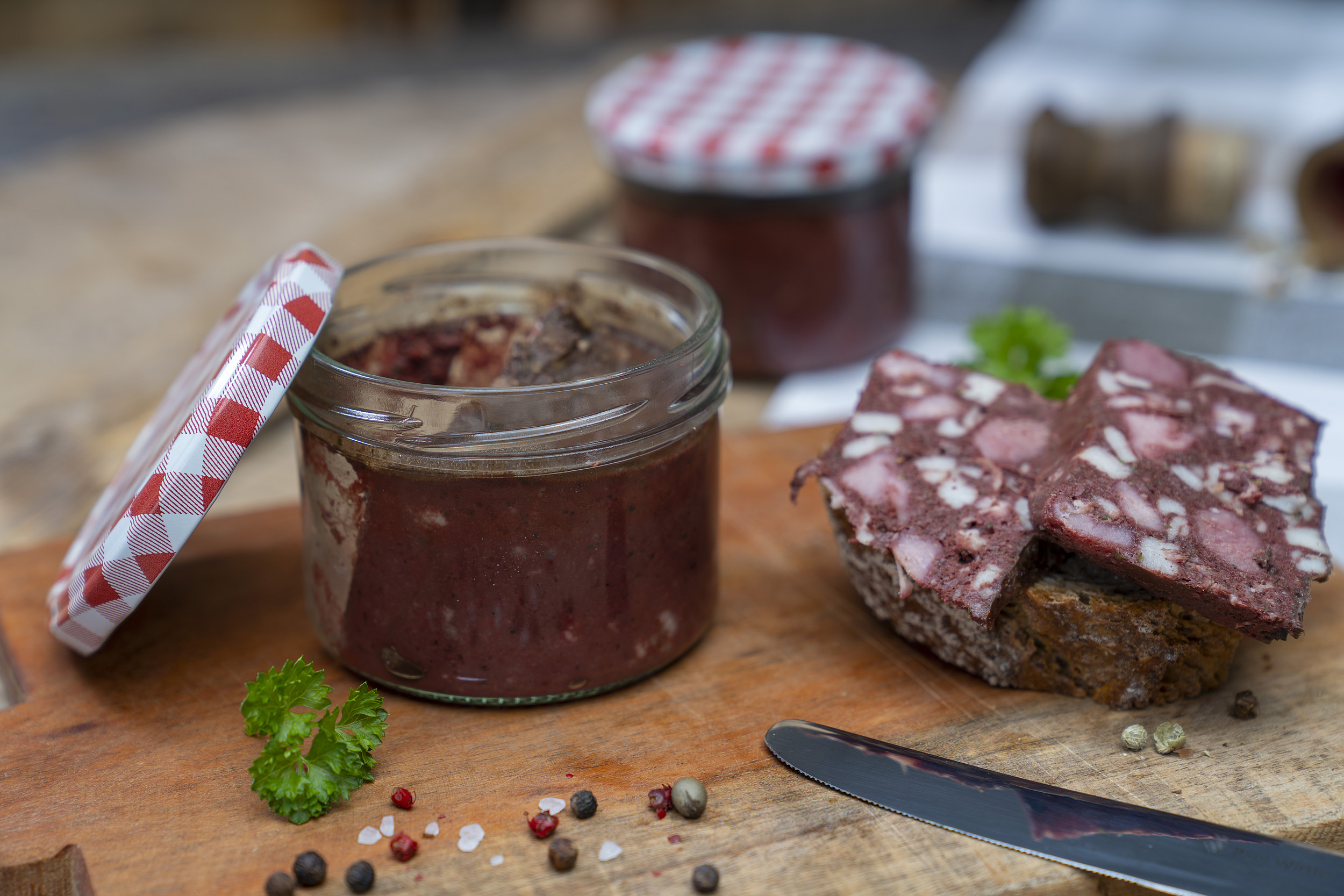 Auf einem Holzbrett steht ein Glas Rotwurst. Daneben eine Scheibe Brot mit Rotwurst Scheiben. Darum Gewürze. Hinten ein weiteres Glas Rotwurst.