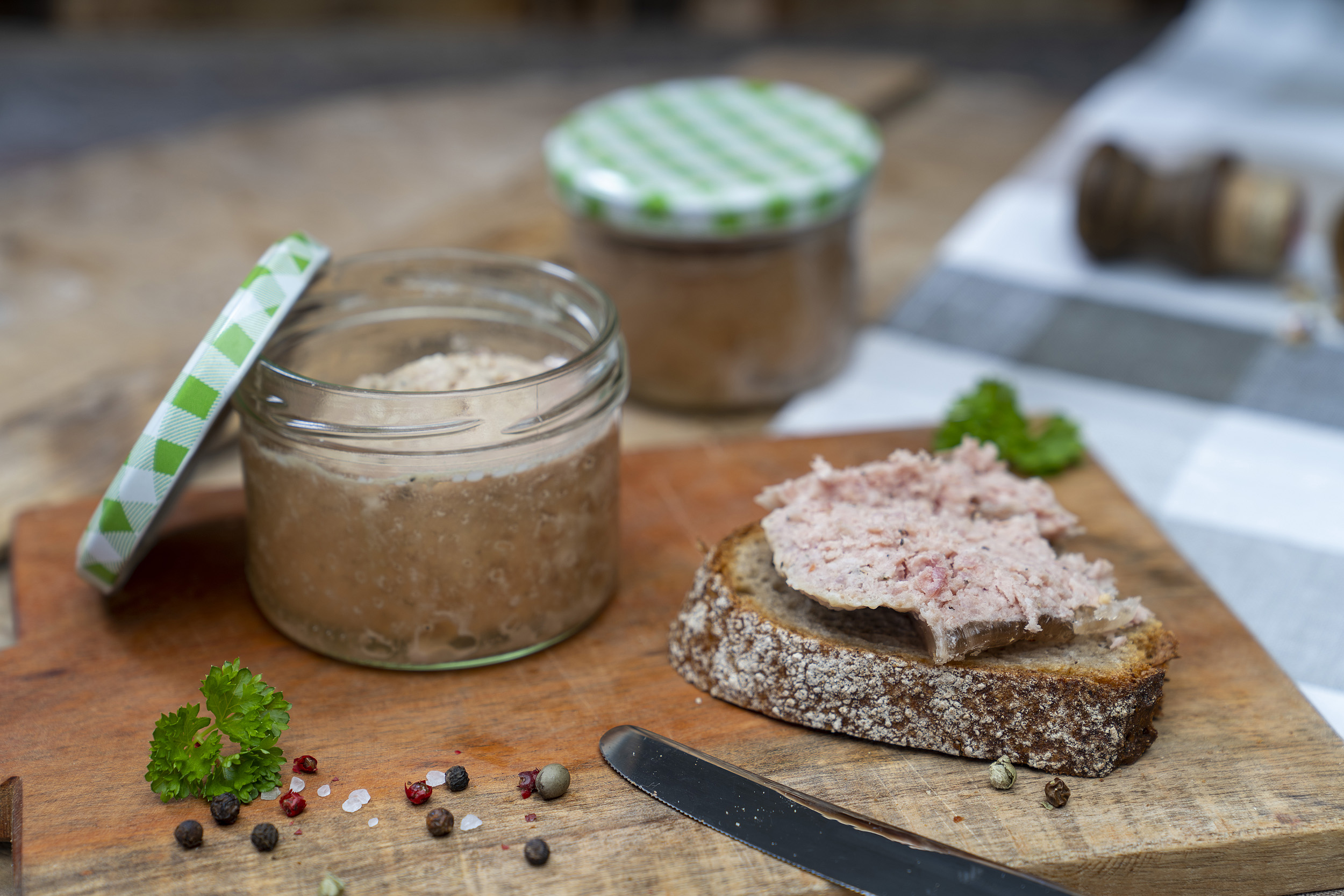 Auf einem Holzbrett steht ein Glas Schartenwurst. Daneben eine Scheibe Brot mit der Wurst. Drum herum Kräuter und Gewürze. Hinten ein weiteres Glas.
