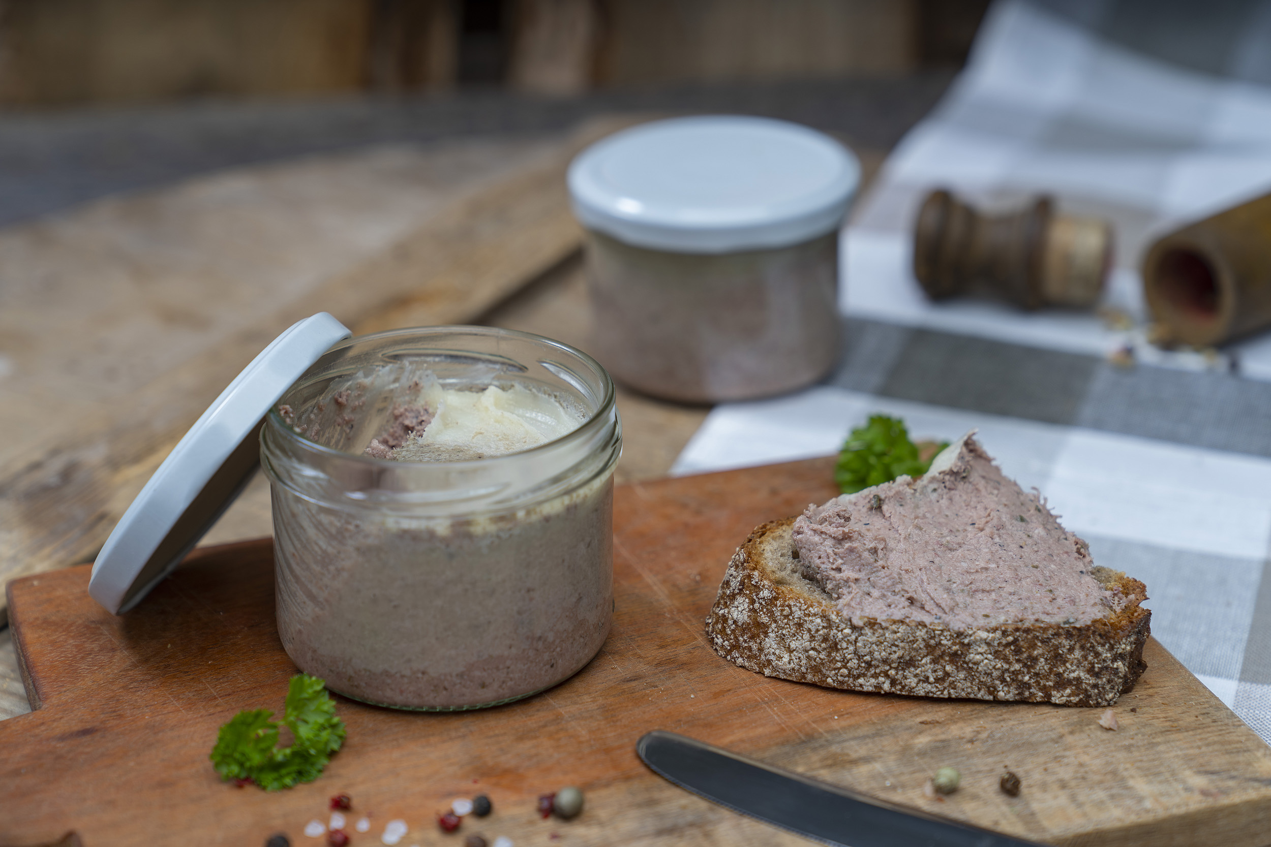 Auf einem Holzbrett ein Glas mit Hausmacher Leberwurst. Daneben eine geschmierte Schnitte, Kräuter und Gewürze. Am Brett ein Messer und im Hintergrund ein weiteres Glas.