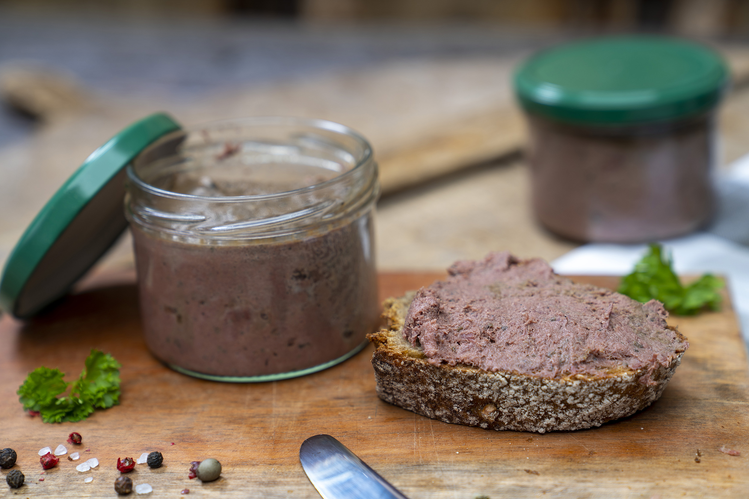 Auf einem Brett ein Glas mit Wildleberwurst. Daneben ein geschmiertes Brot und Gewürze. Im Hintergrund noch ein Glas.