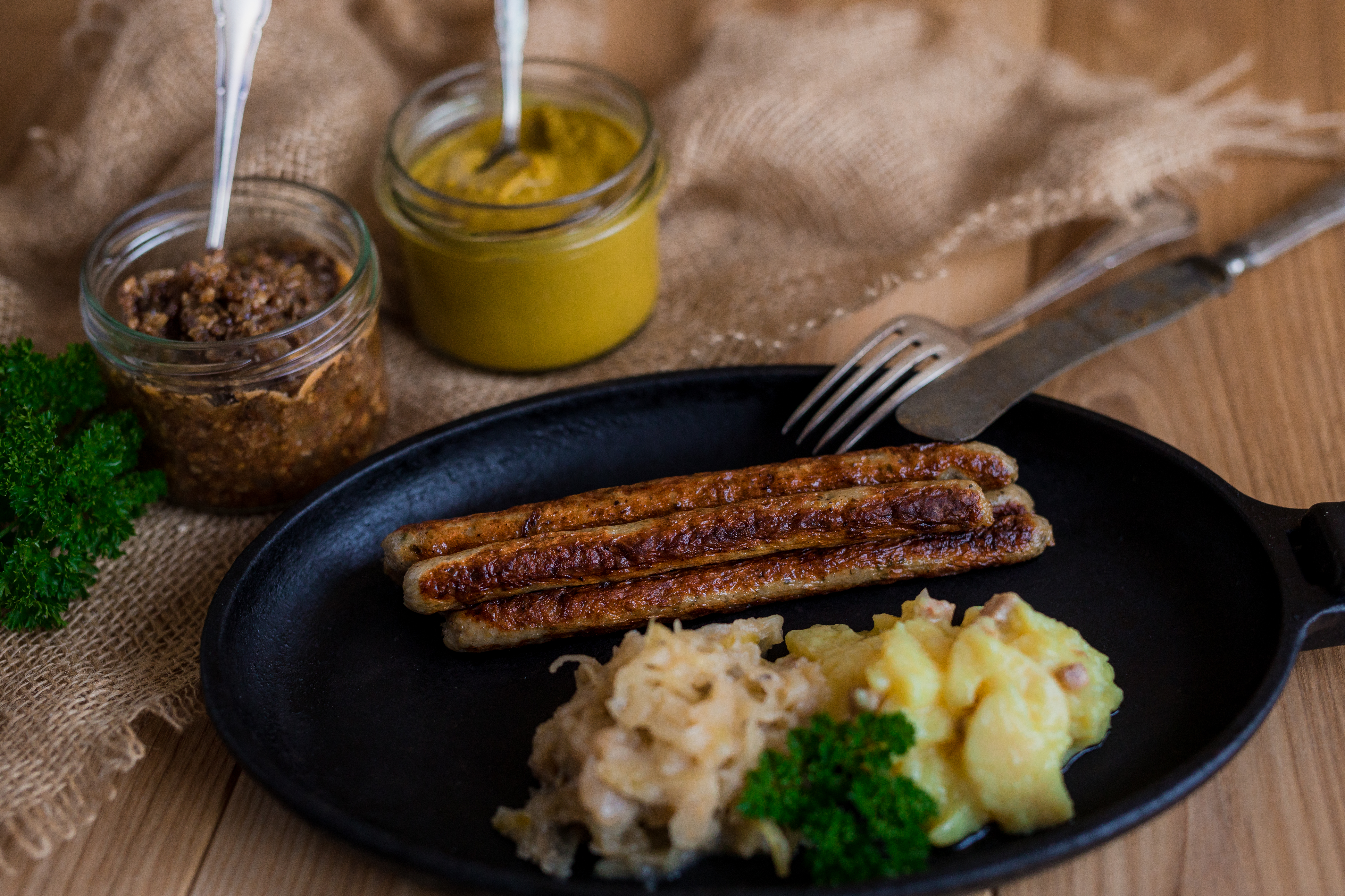 Gebratene Klammerwürste in einer Pfanne mit Sauerkraut und Salatbeilage. Neben der Pfanne Gläser mit Saucen. Untergrund aus Holz.