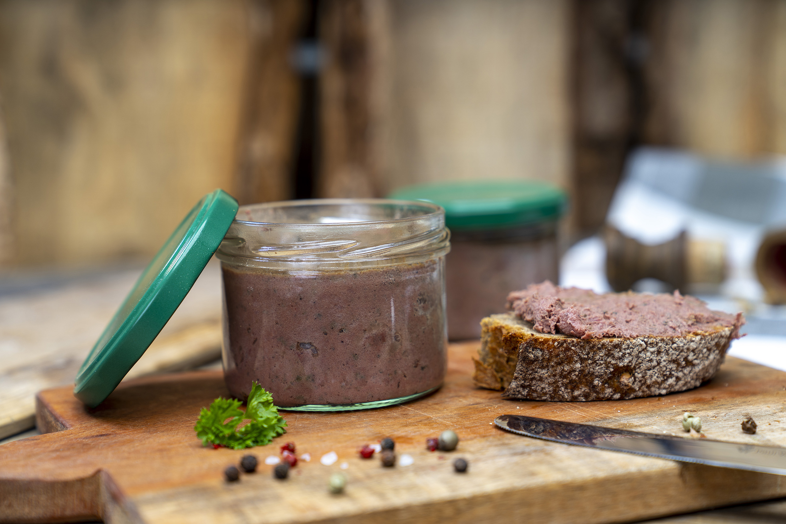 Auf einem Brett ein Glas mit Wildleberwurst. Daneben ein geschmiertes Brot und Gewürze. Im Hintergrund noch ein Glas.