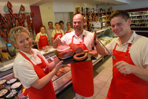 Bettina, Erwin und Hendrik Fessel mit roten Schürzen im Vordergrund. Neueröffnung des Ladens in 2010. Im Hintergrund die Theke und das Team.