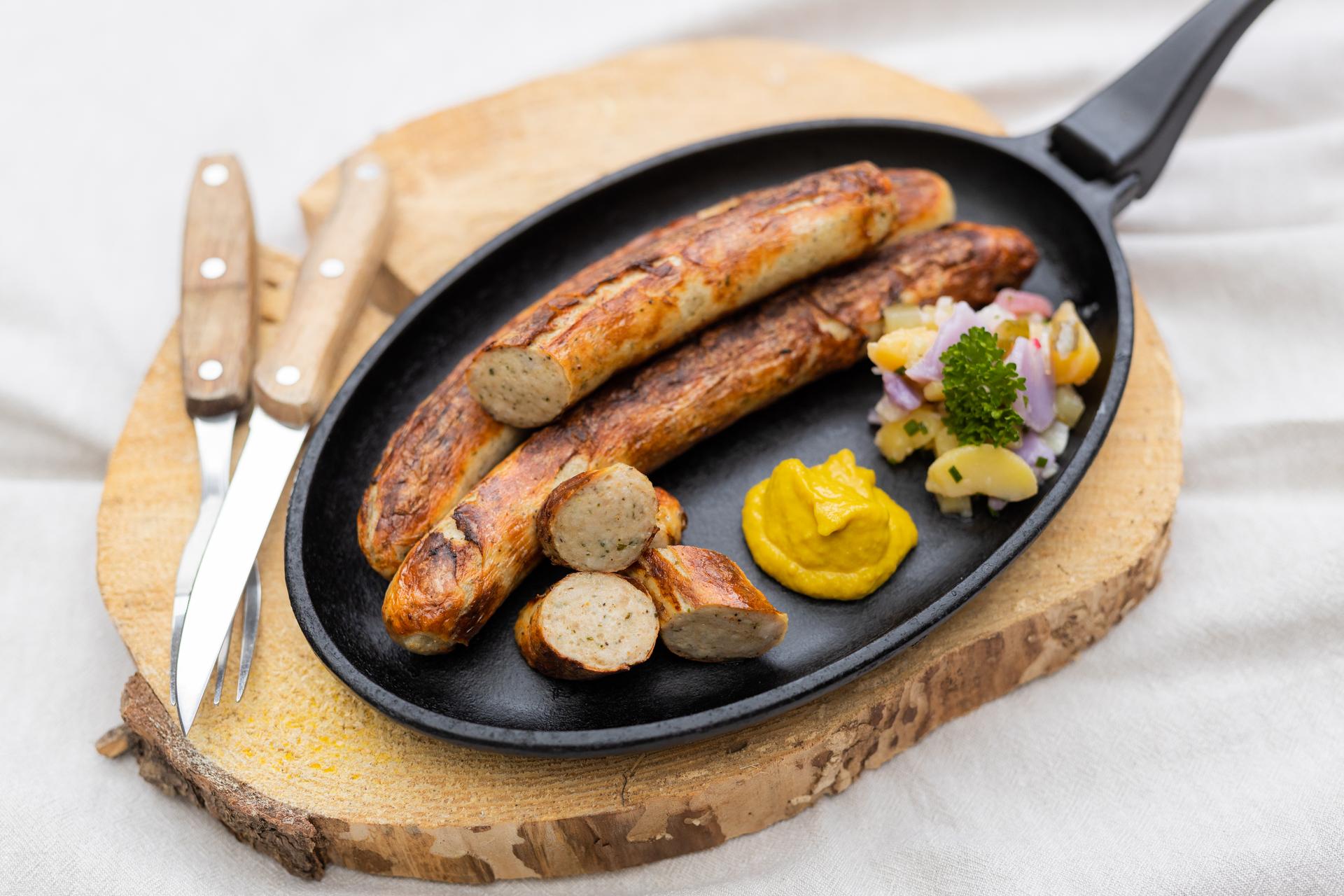 Gebratene Original Thüringer Rostbratwurst in der Pfanne auf einem Brett. In der Pfanne Senf und Salatbeilage. Neben der Pfanne Besteck.