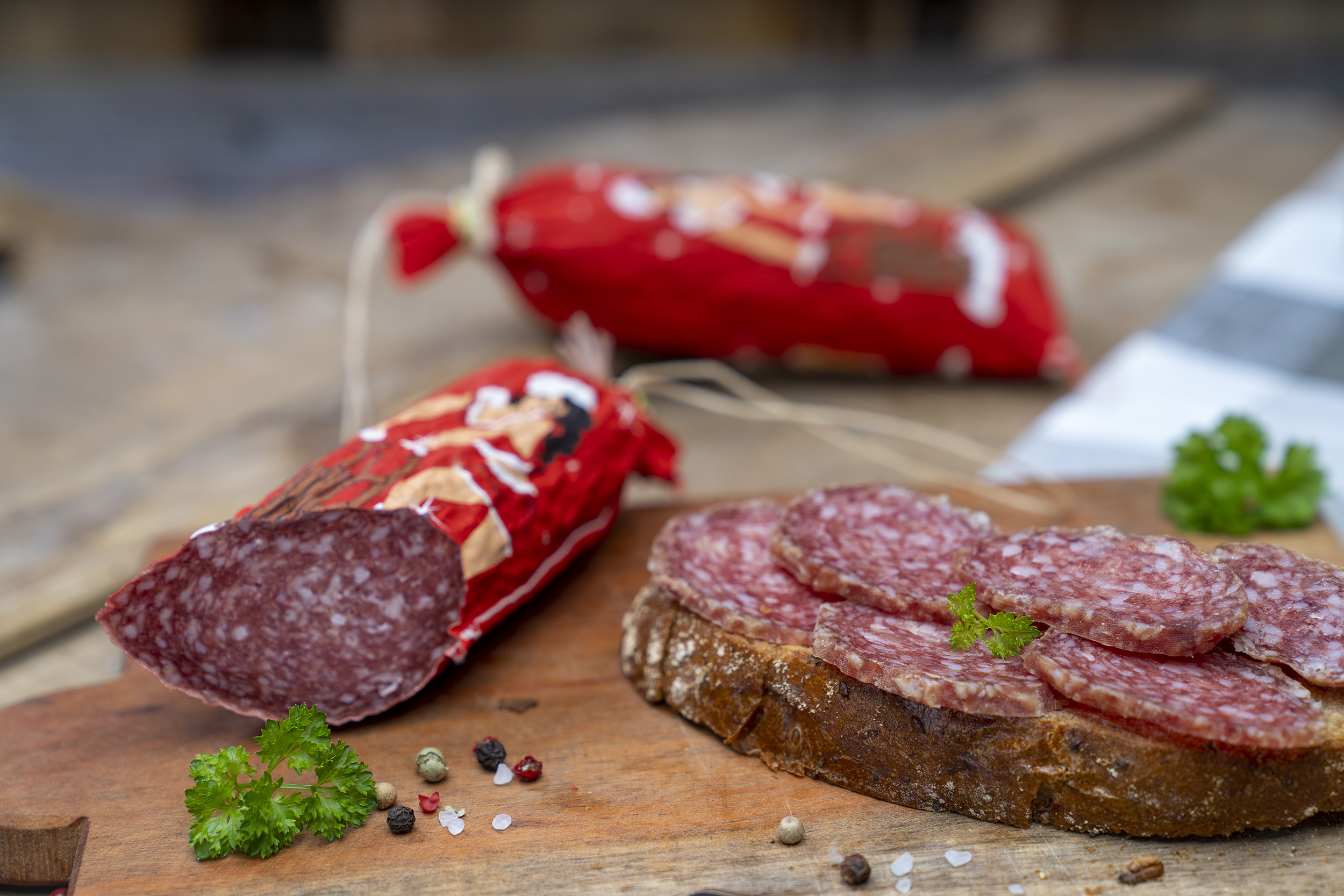 Salami mit weihnachtlichen Wurstmantel auf einem Brett. Daneben eine Brotscheibe mit der Wurst und Gewürze. Im Hintergrund eine weitere Salami.