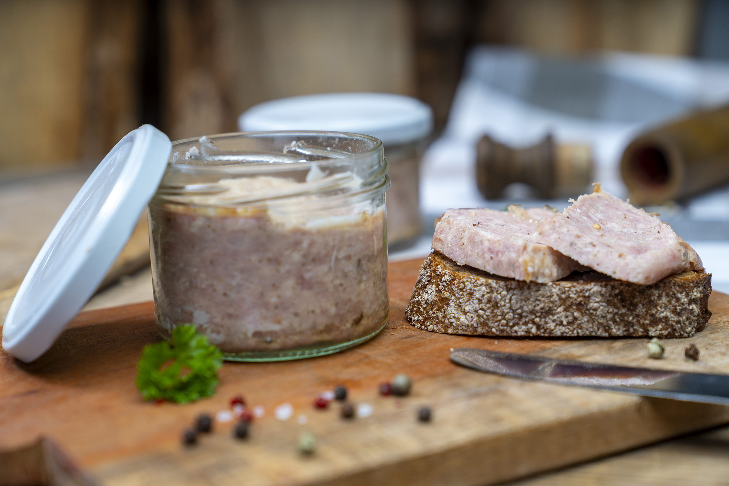 Auf einem Holzbrett ein Glas mit Mettwurst. Daneben eine Schnitte mit Mettwurst, Kräuter und Gewürze. Im Hintergrund steht ein weiteres Glas. Vorn lehnt ein Messer am Brett.