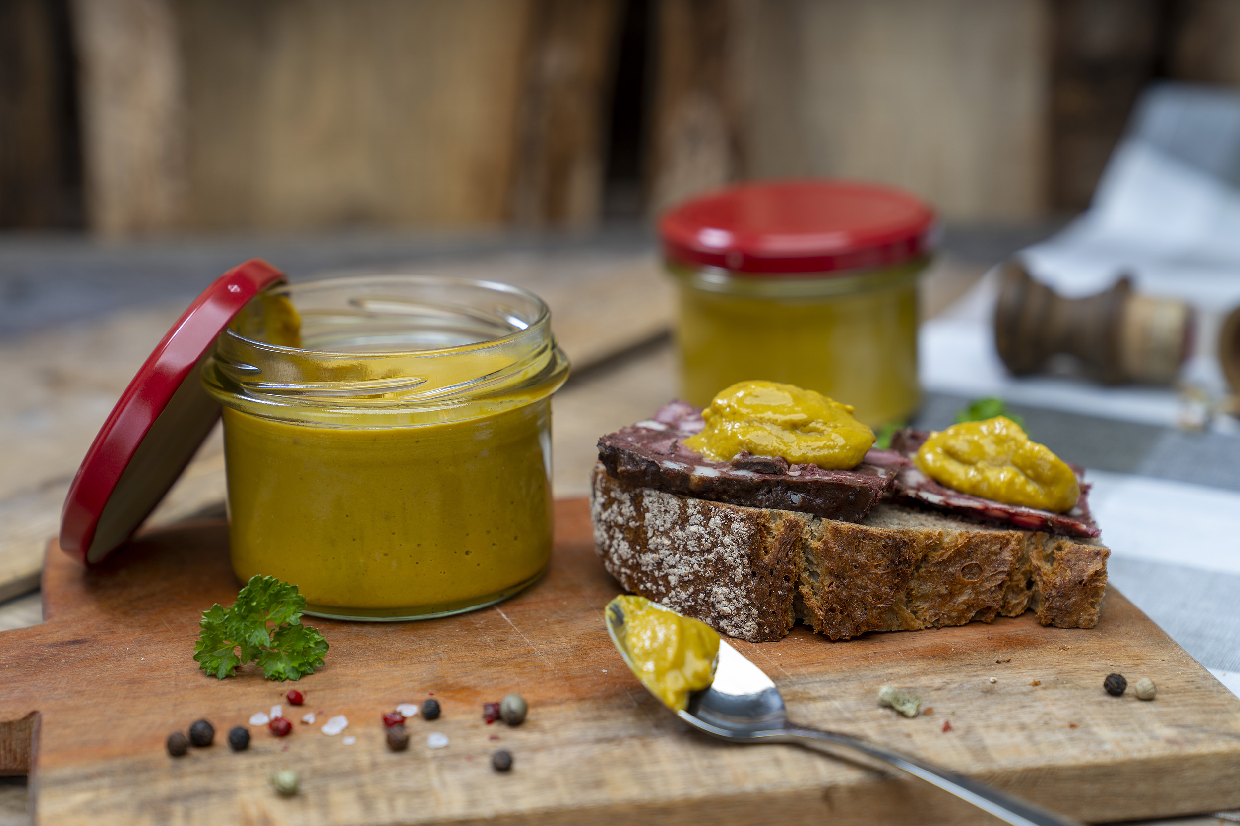 Auf einem Holzbrett ein Glas Senf bzw. Meisters Mostrich. Daneben eine Schnitte mit Rotwurst und Senf. Darum Gewürze und Kräuter. Vorn ein Löffel mit etwas Senf. Hinten ein weiteres Glas.