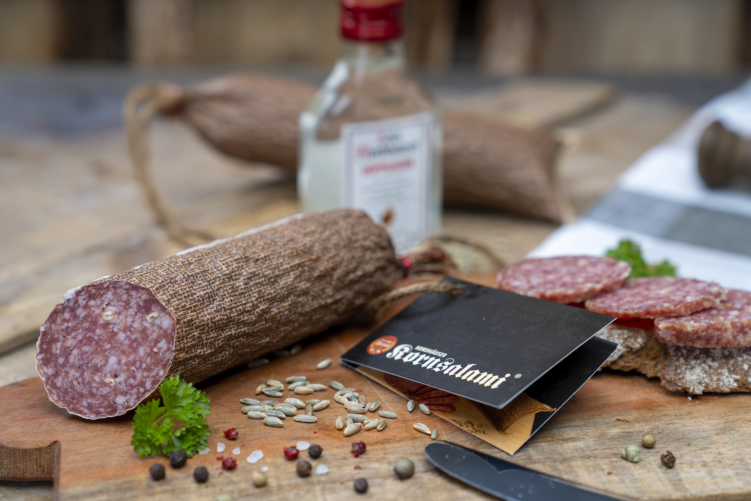 Kornsalami auf einem Holzbrett. Daneben eine Scheibe Brot mit der Salami und Gewürze. Im Hintergrund eine kleine Flasche Echter Nordhäuser Doppelkorn.