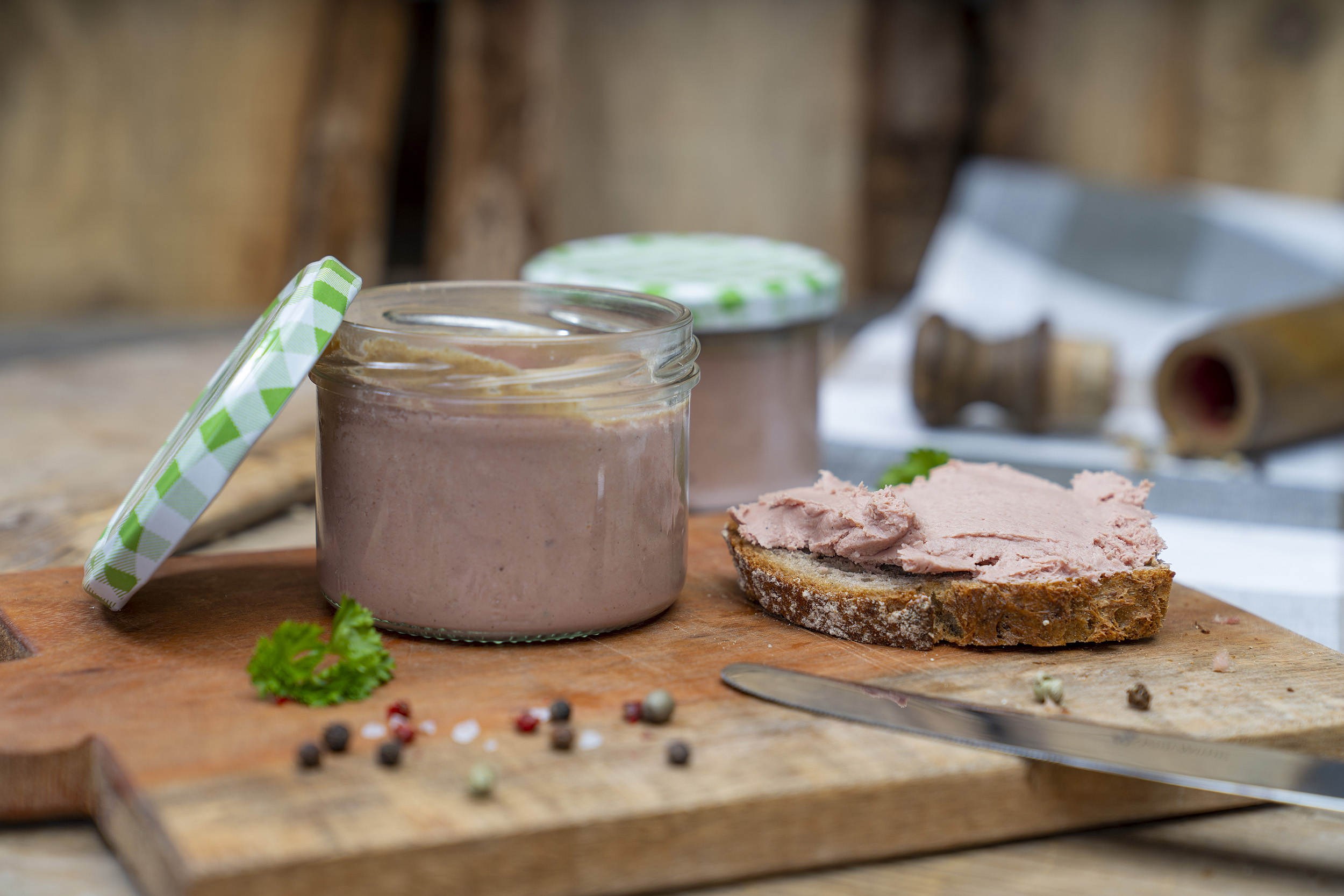 Auf einem Holzbrett ein Glas mit Kalbfleischleberwurst. Daneben eine Schnitte mit Leberwurst beschmiert und Gewürze. Am Brett vorn liegt ein Messer. Im Hintergrund ein weiteres Glas.