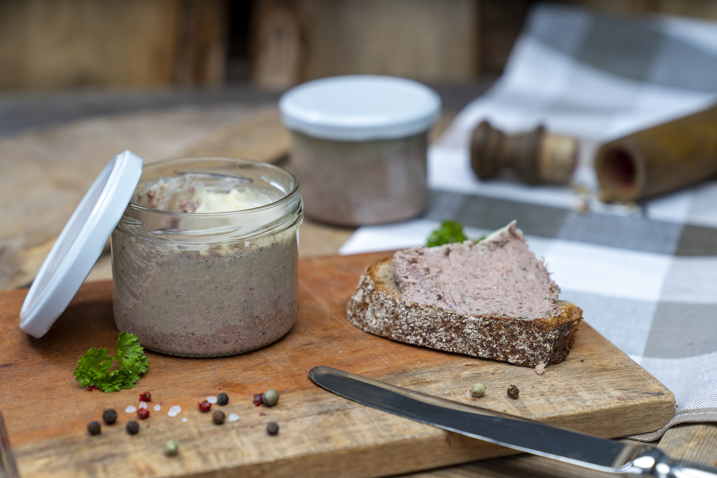 Auf einem Holzbrett ein Glas mit Hausmacher Leberwurst. Daneben eine geschmierte Schnitte, Kräuter und Gewürze. Am Brett ein Messer und im Hintergrund ein weiteres Glas.