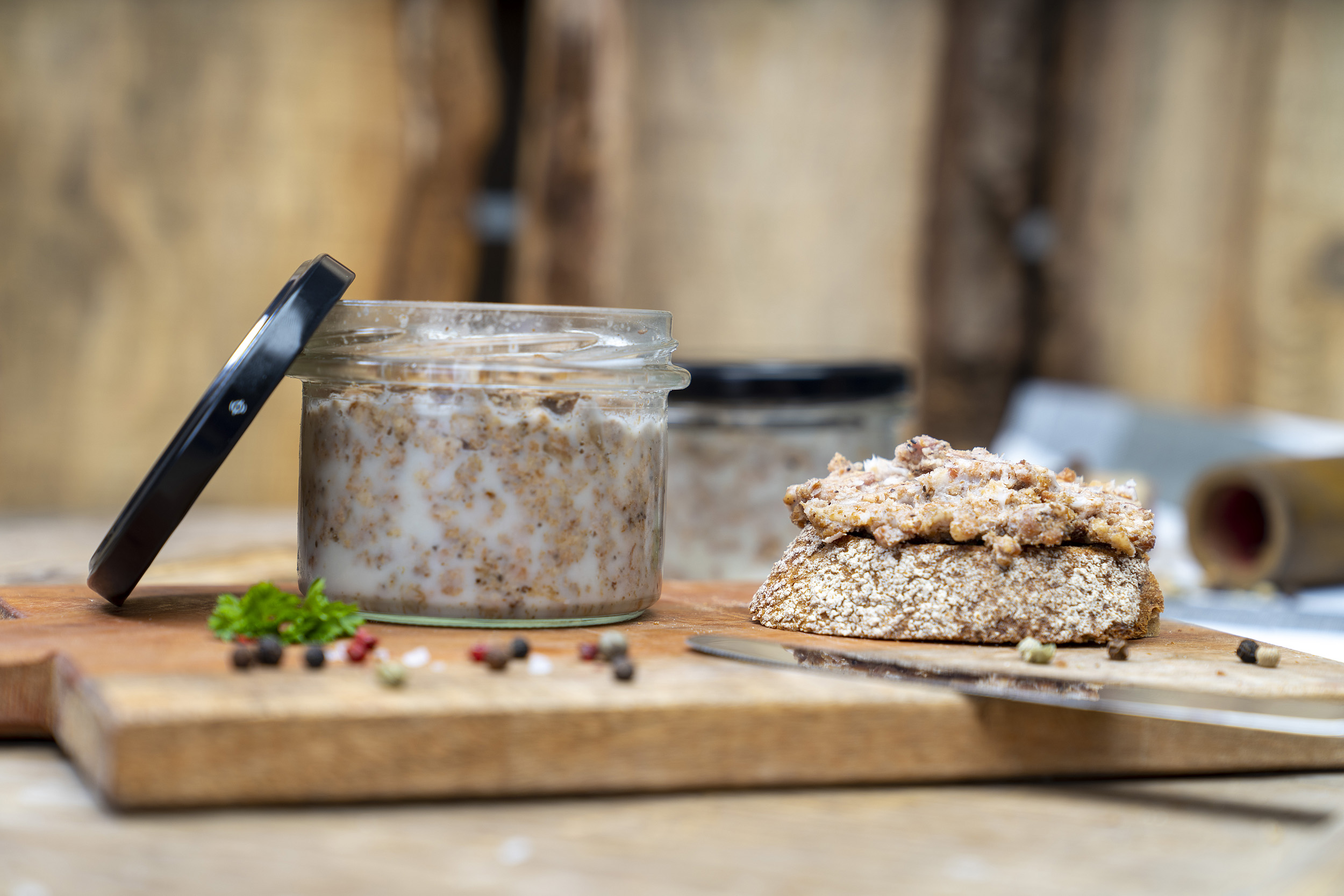 Auf einem Holzbrett ein Glas Pott Suse, also geschmortes Hackfleisch. Neben dem Glas eine geschmierte Schnitte und Gewürze. Im Hintergrund ein weiteres Glas.