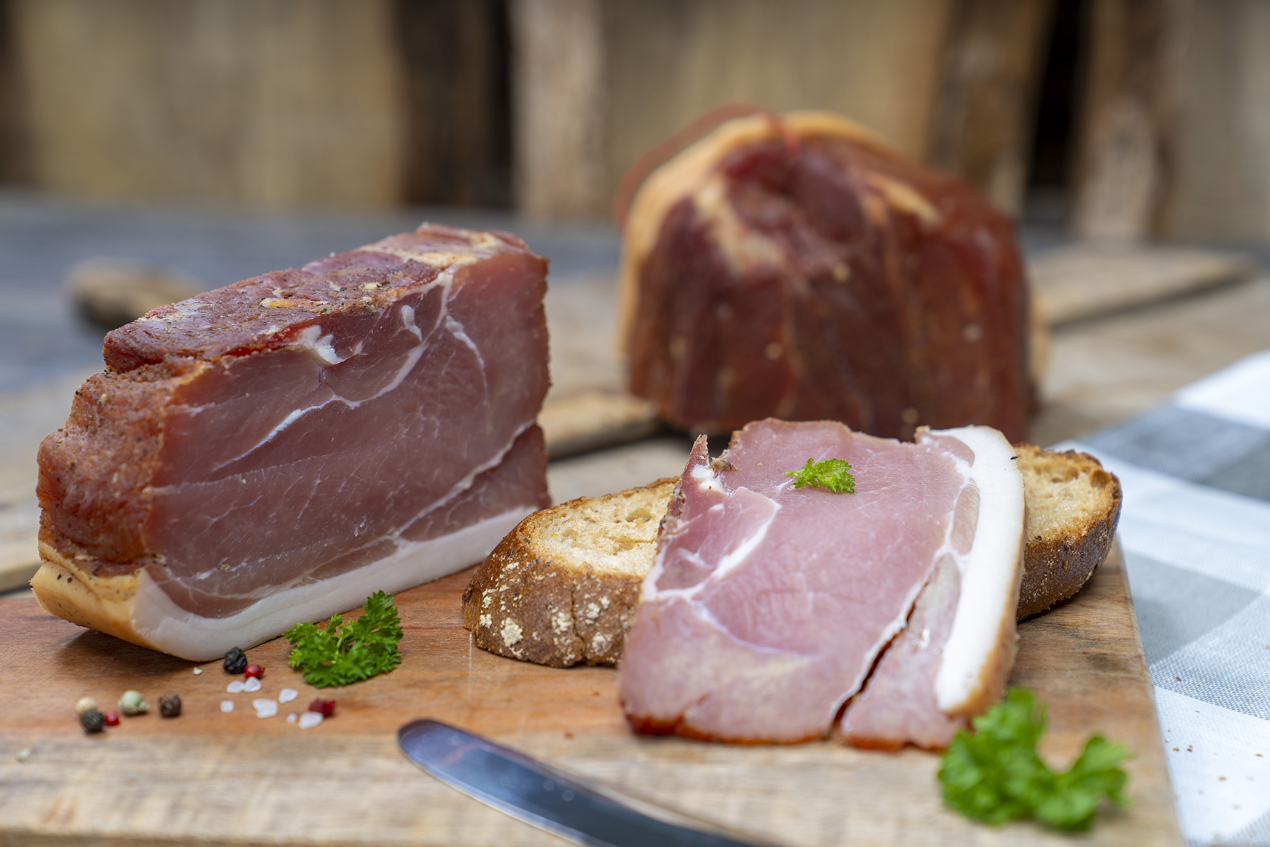 Auf einem Brot eine Scheiben Schinkenspeck. Daneben der Schinken und Gewürze. Im Hintergrund ein weiterer Schinken der Fleischerei Fessel.