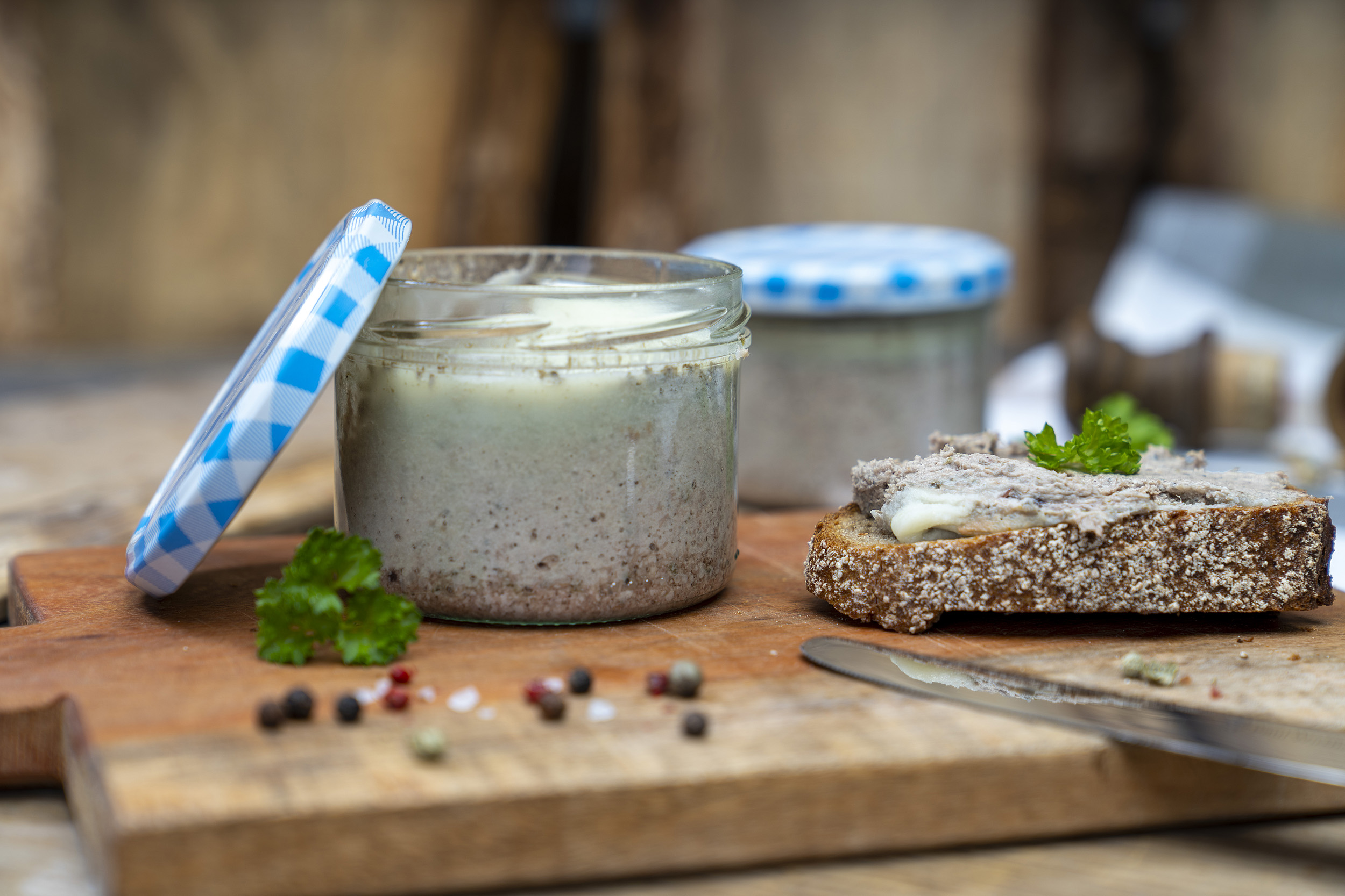 Ein Glas Zwiebelleberwurst auf eine Brett. Ein beschmiertes Brot und Gewürze daneben. Hinten ein weiteres Glas der Leberwurst.
