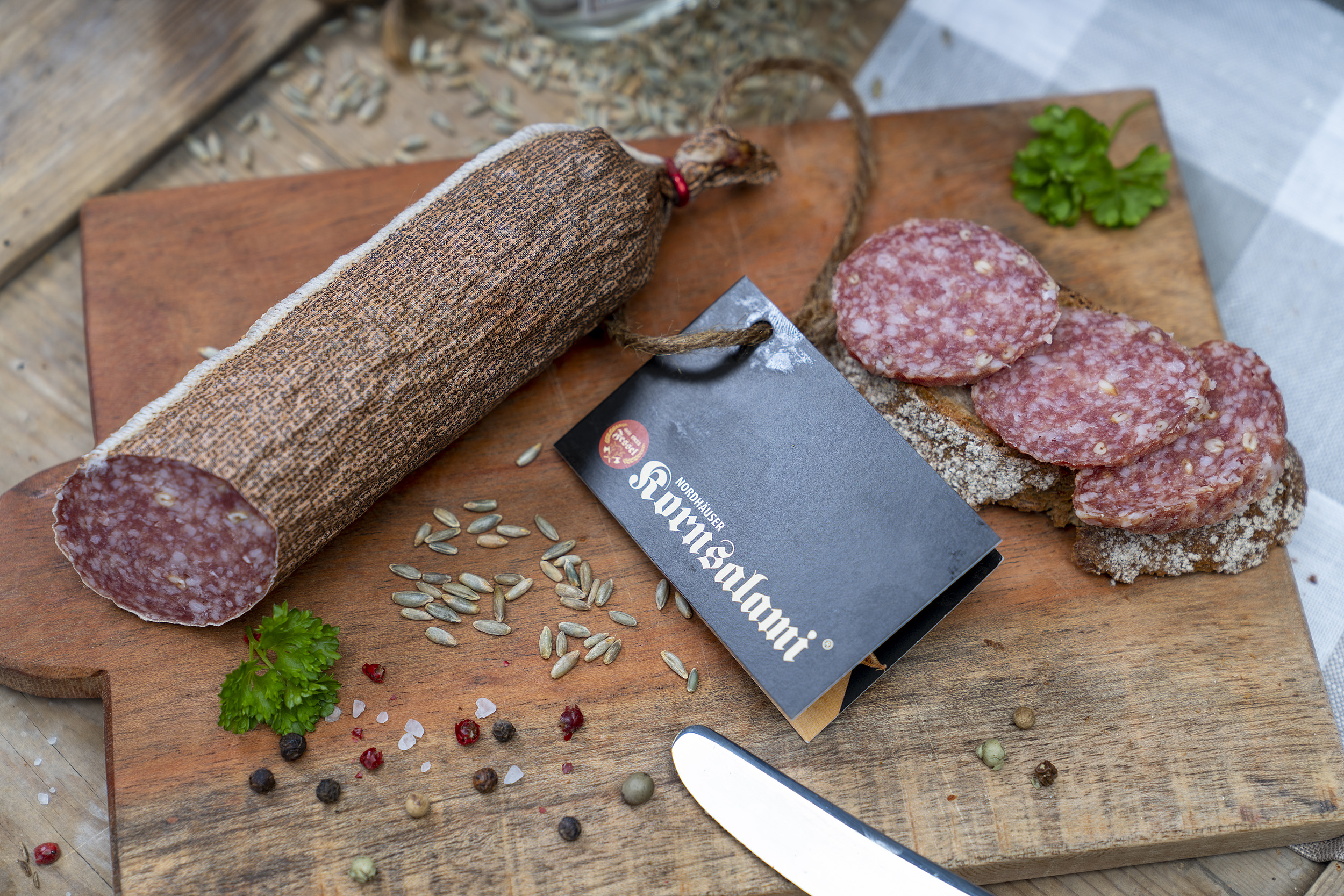 Kornsalami auf einem Holzbrett. Daneben eine Scheibe Brot mit der Salami und Gewürze.