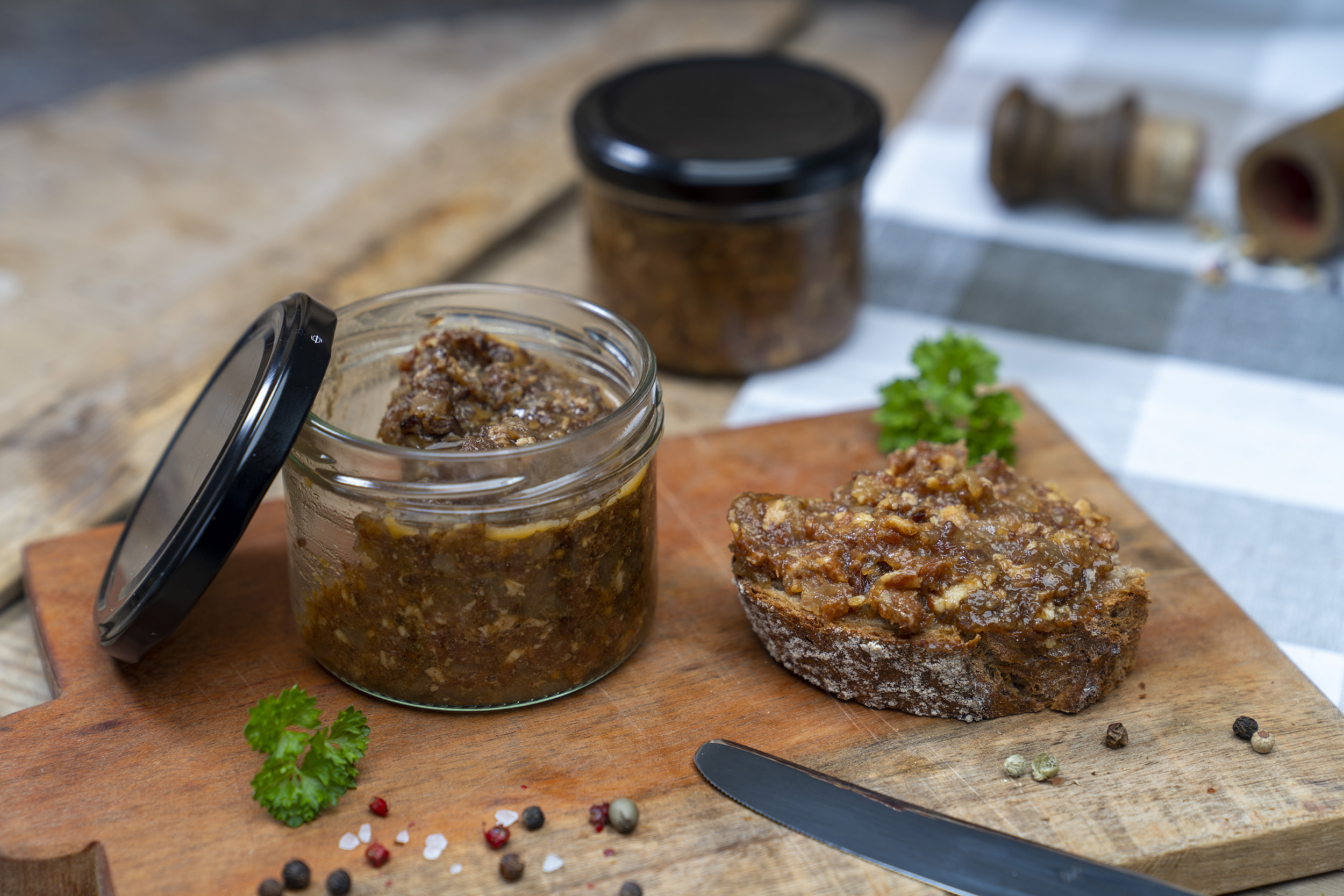 Auf einem Holzbrett steht ein Glas mit Bacon Jam. Daneben Gewürze, Kräuter und ein geschmiertes Brot mit der Marmelade. Im Hintergrund ein weiteres Glas. Vorn ein Messer am Brett.