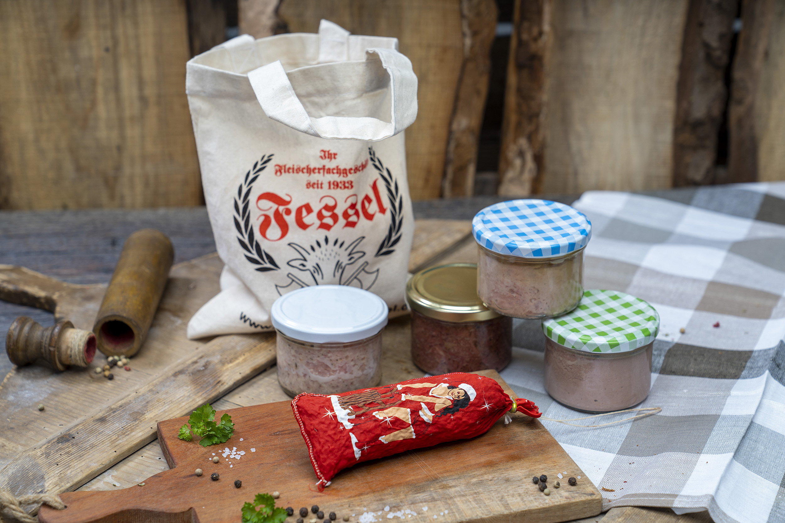 Auf einem Tisch vier verschiedene Konsevengläser Wurstprodukte und eine Salami in weihnachtlichem Look. Hinten ein Jutebeutel der Fleischerei Fessel.