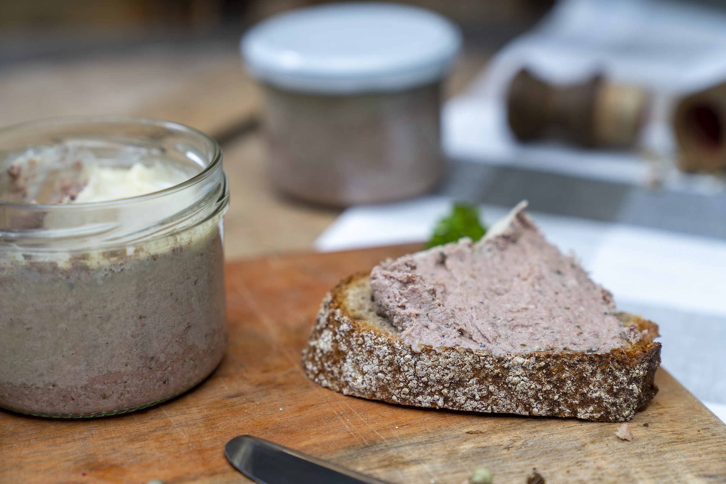 Auf einem Holzbrett ein Glas mit Hausmacher Leberwurst. Daneben eine geschmierte Schnitte, Kräuter und Gewürze. Am Brett ein Messer und im Hintergrund ein weiteres Glas.