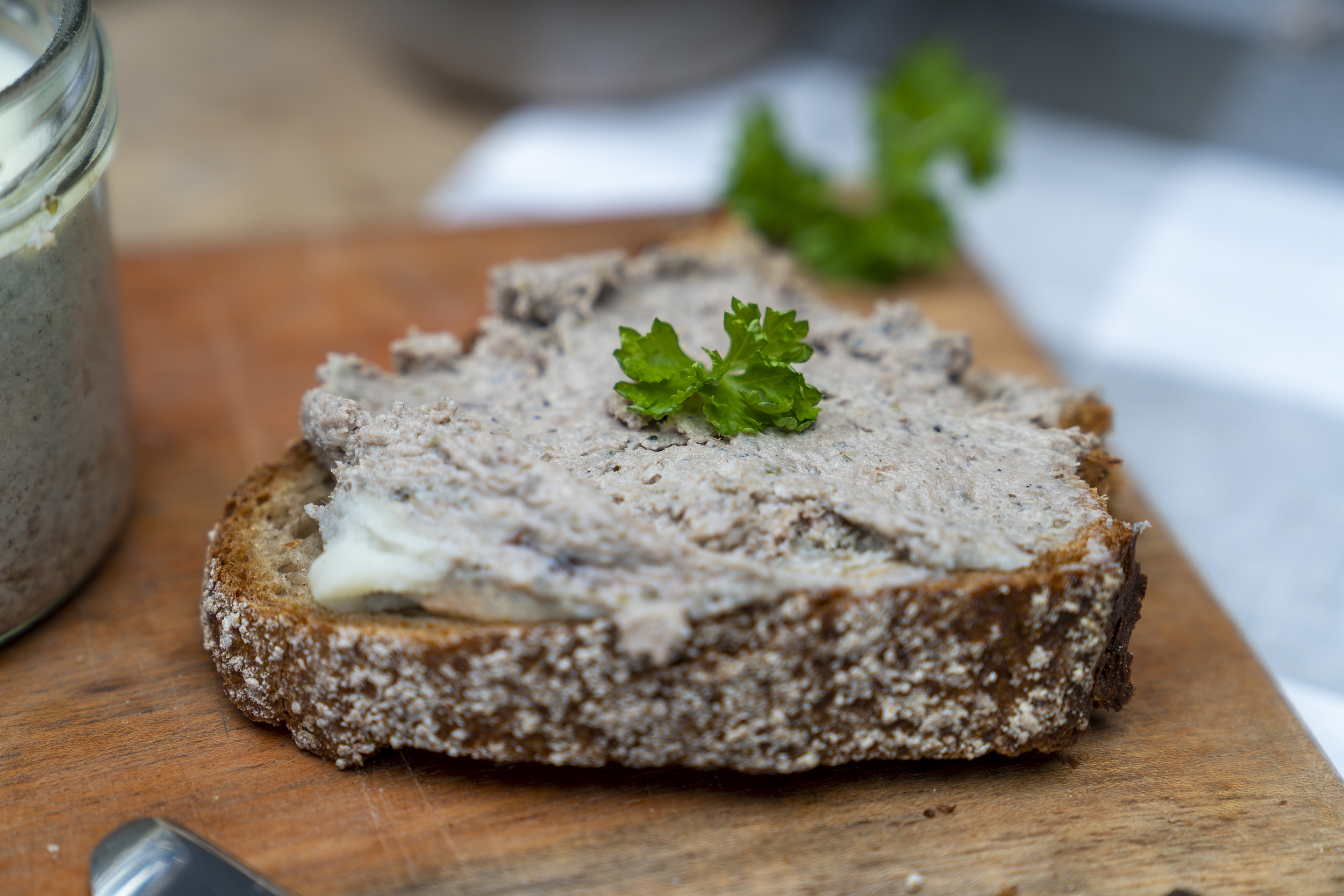 Ein geschmiertes Brot mit Leberwurst auf einem Brett. Darauf Petersilie und daneben das geöffnete Glas.