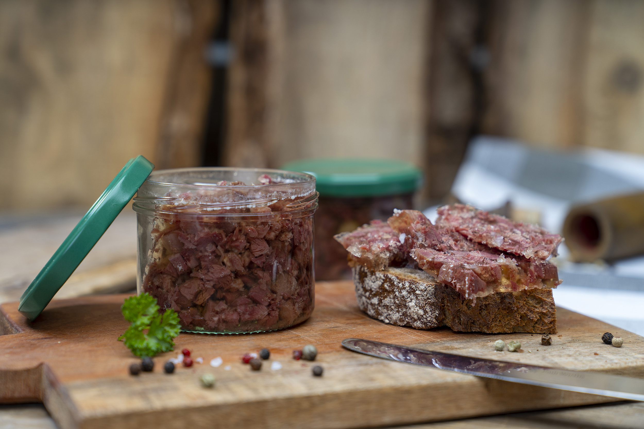 Ein Glas Wildsuelze auf einem Brett. Daneben ein geschmiertes Brot und Gewürze. Hinten ein weiteres Glas.