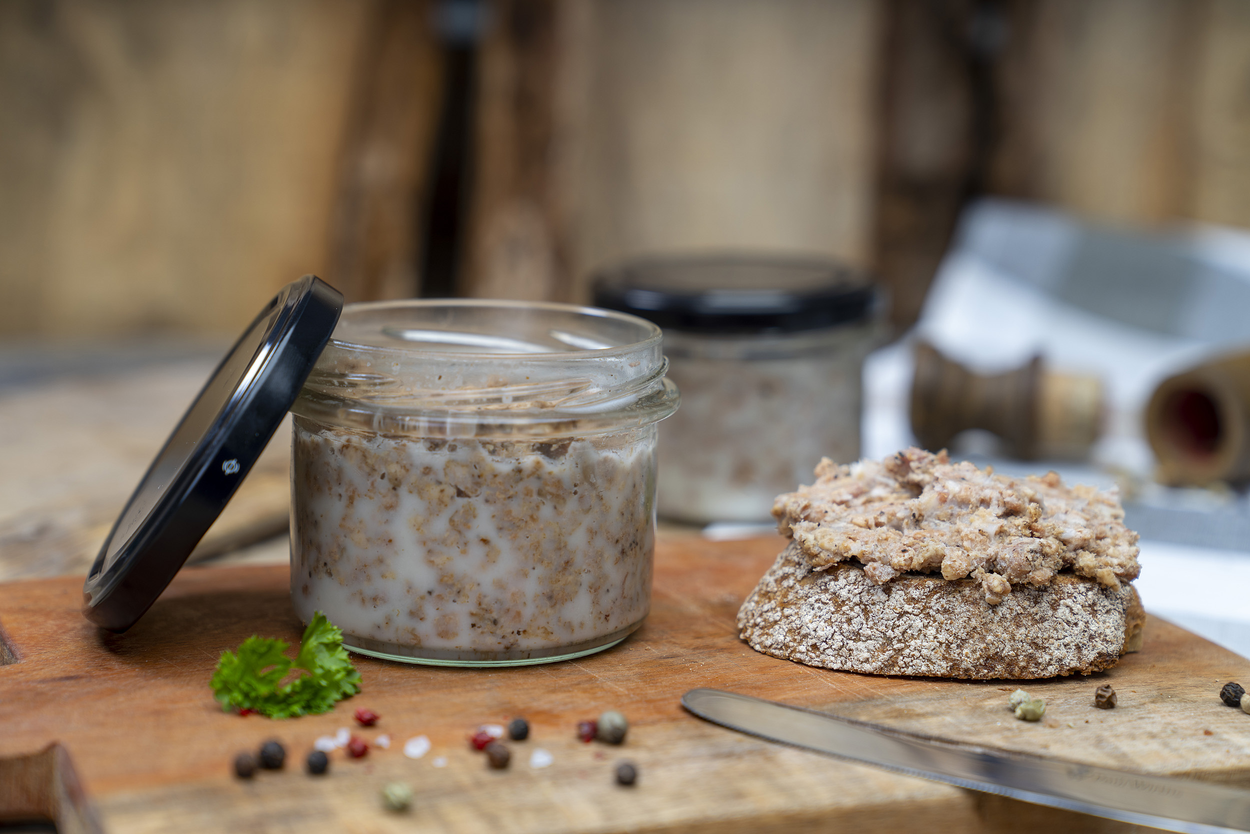 Auf einem Holzbrett ein Glas Pott Suse, also geschmortes Hackfleisch. Neben dem Glas eine geschmierte Schnitte und Gewürze. Im Hintergrund ein weiteres Glas.