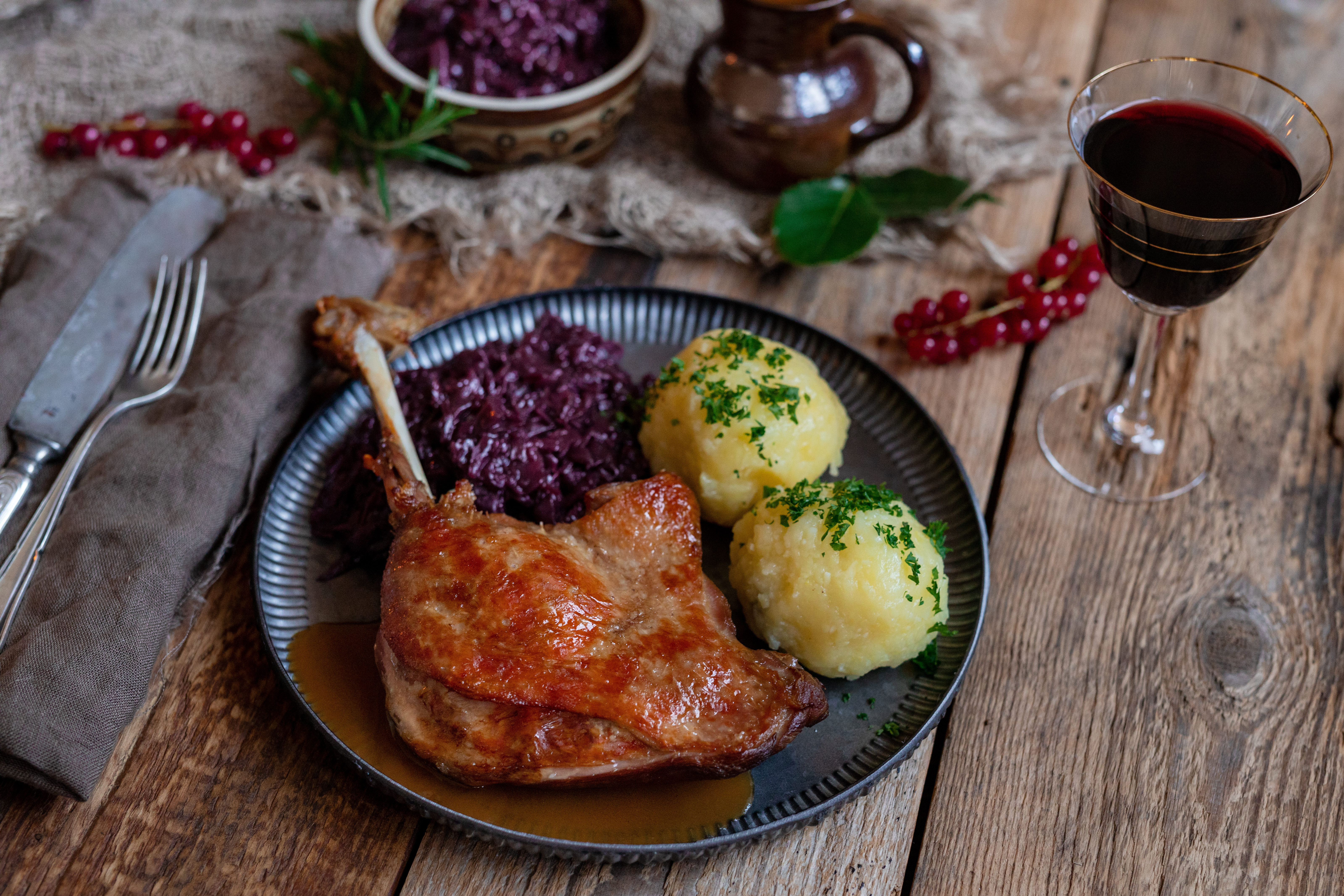 Gänsekeule, Rotkohl und Klöße auf einem Tisch. Daneben Besteck, ein Glas Rotwein und Deko.