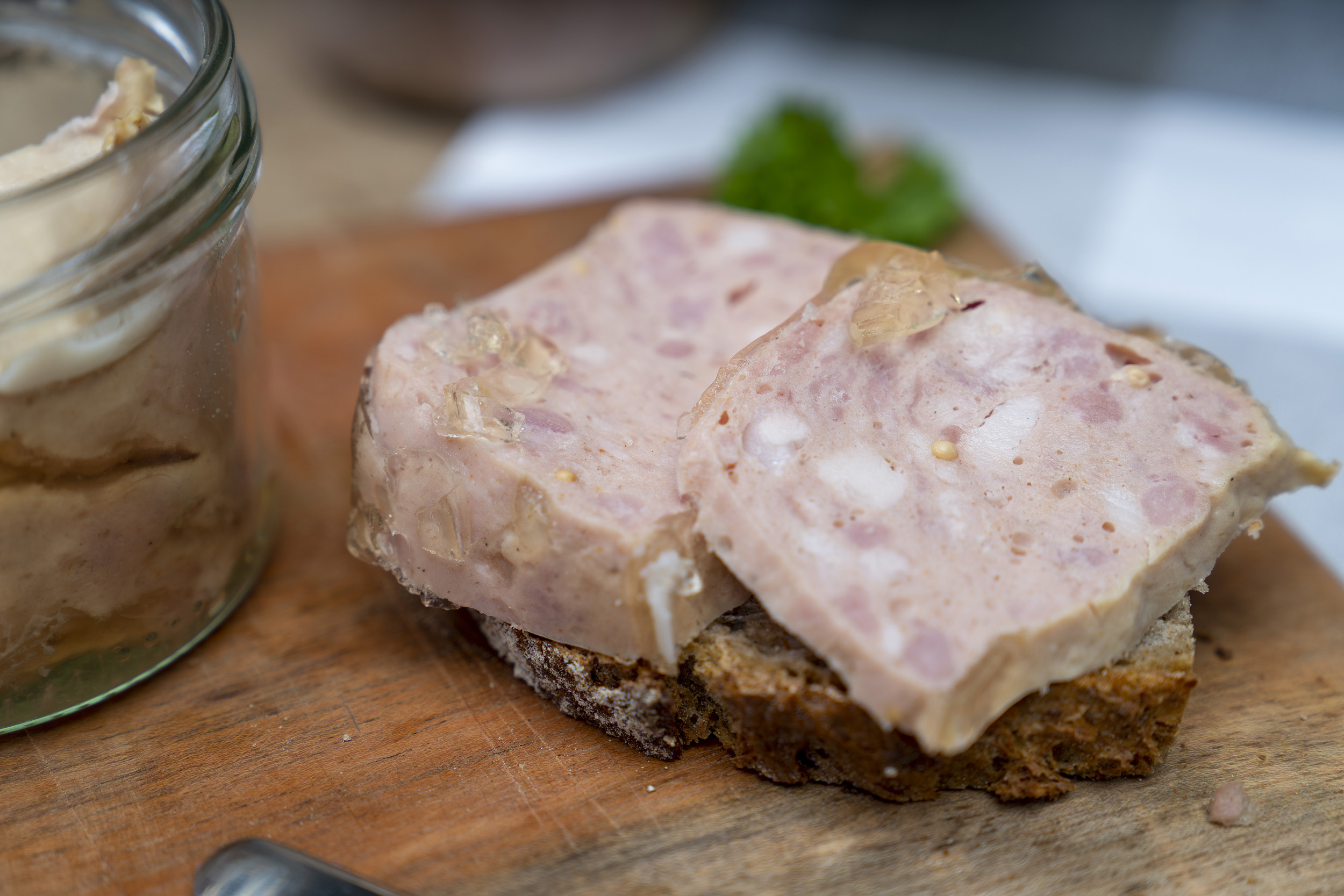 Nahaufnahme von einer Schnitte mit Jagdwurst auf einem Holzbrett. Daneben ein Glas, hinten Kräuter.