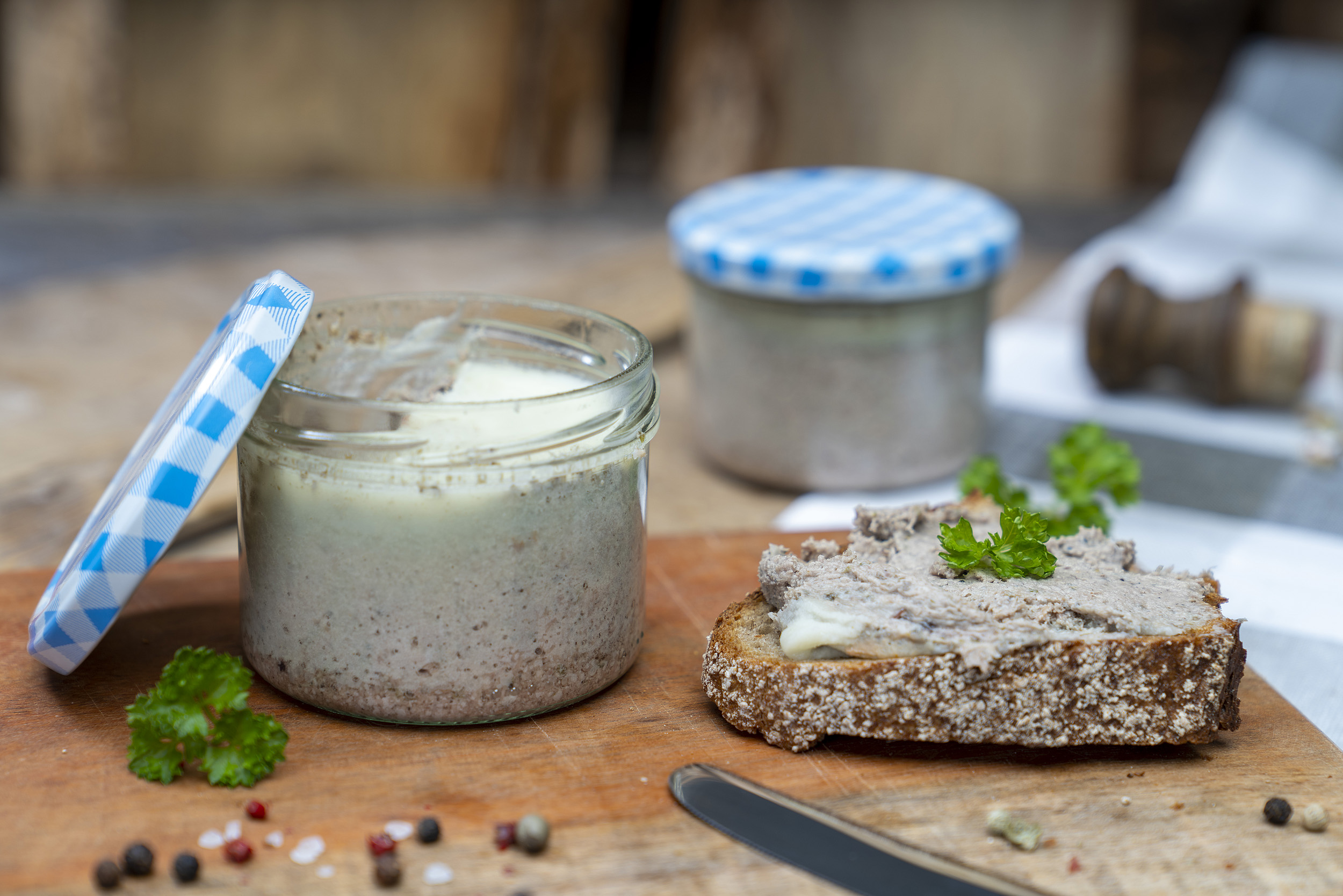 Ein Glas Zwiebelleberwurst auf eine Brett. Ein beschmiertes Brot und Gewürze daneben. Hinten ein weiteres Glas der Leberwurst.