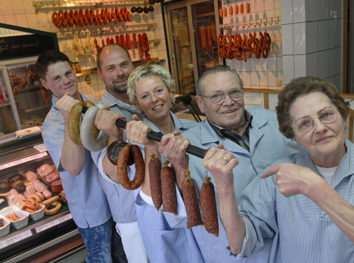 Die Familie Fessel hiintereinander im alten Laden. Sie halten gemeinsam einen Stock mit verschiedenen Würsten. Vorn Anneliese und Gerhard, dann Bettina und Erwin, ganz hinten Hendrik Fessel. Alle tragen blau-weiße Kittel. Im Hintergrund die Wursttheke.
