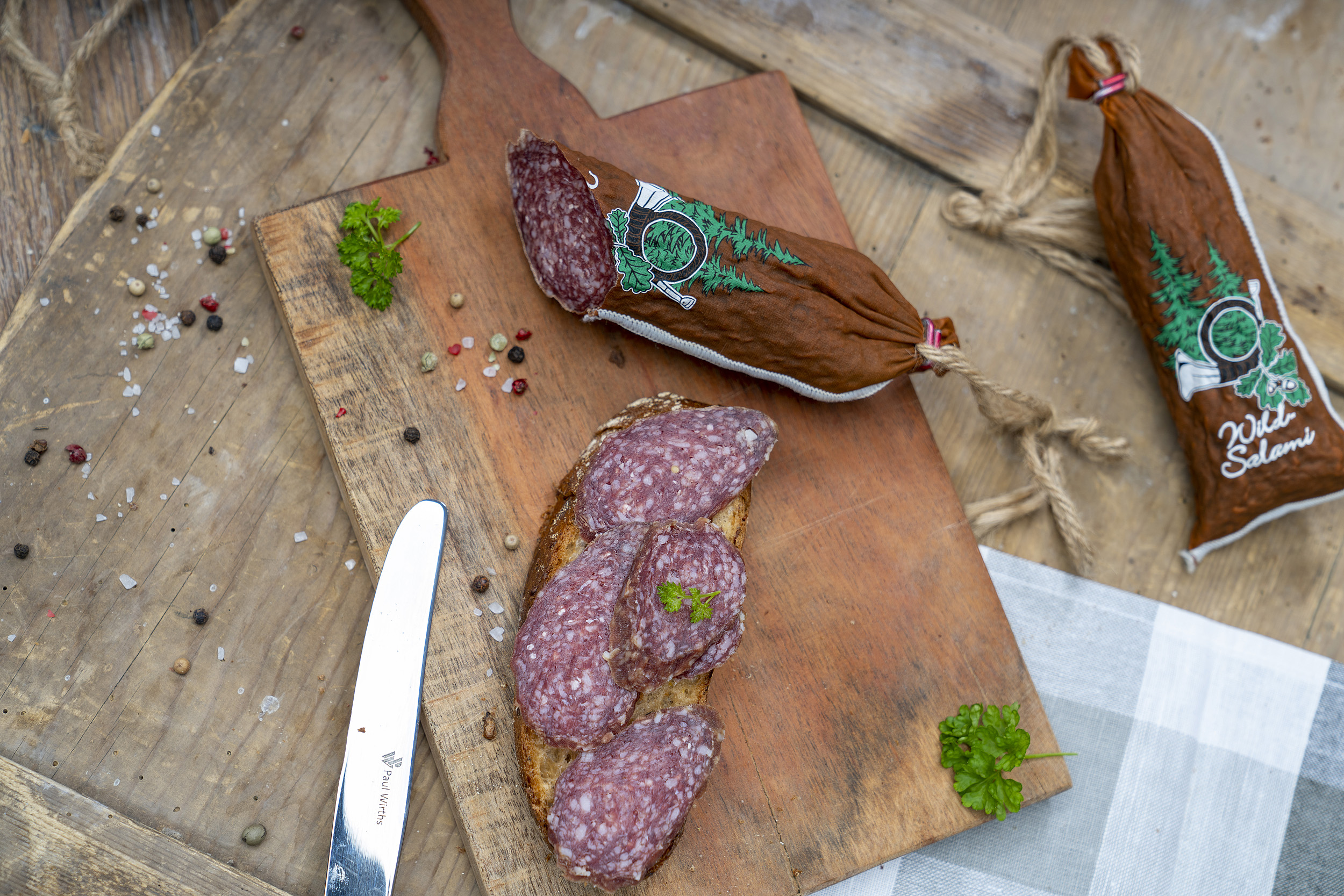 Hirschsalami auf Holzbrett. Daneben eine Schnitte mit Salami und Kräuter. Daneben noch eine Hirschsalami.