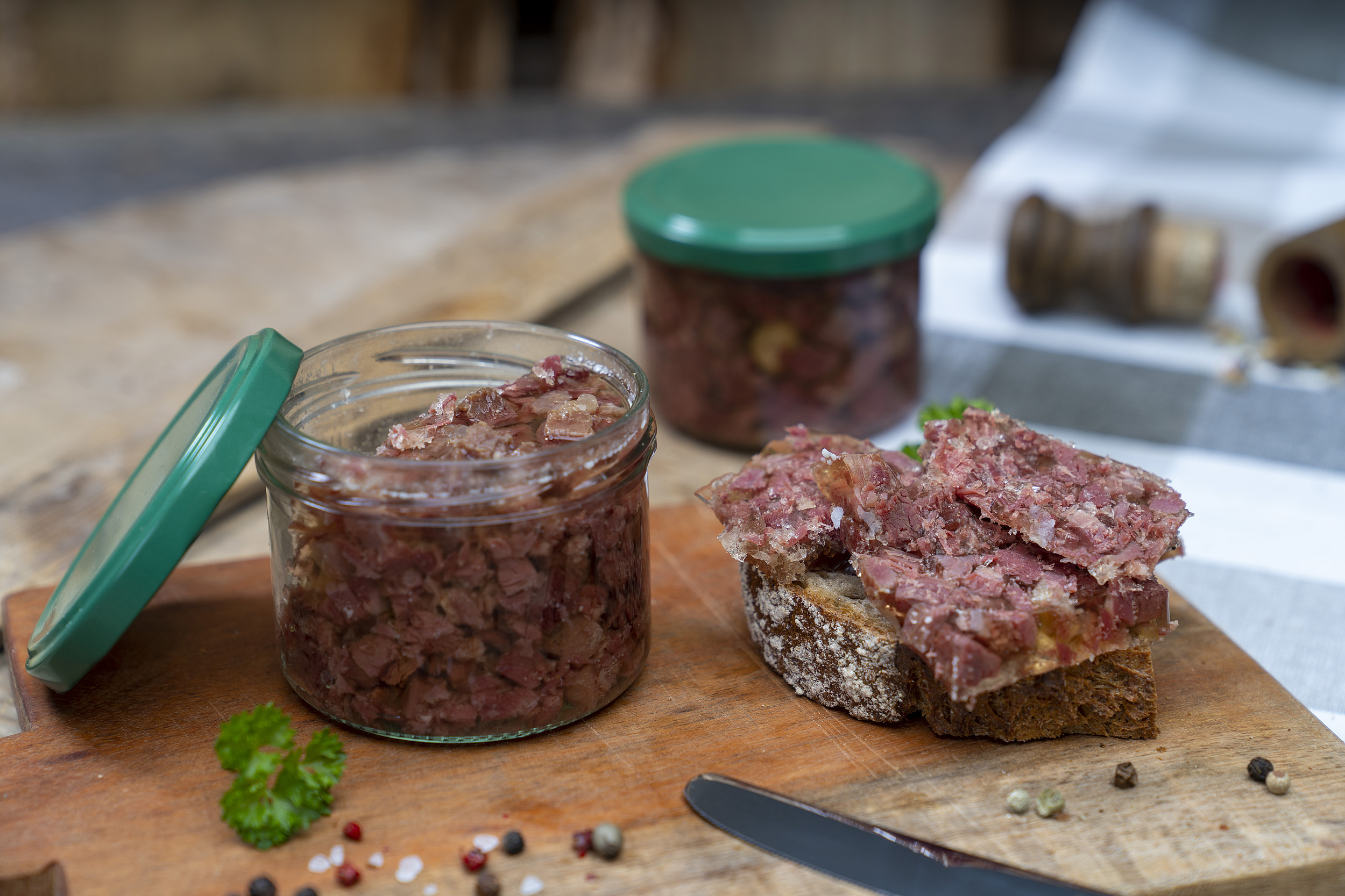 Ein Glas Wildsuelze auf einem Brett. Daneben ein geschmiertes Brot und Gewürze. Hinten ein weiteres Glas.