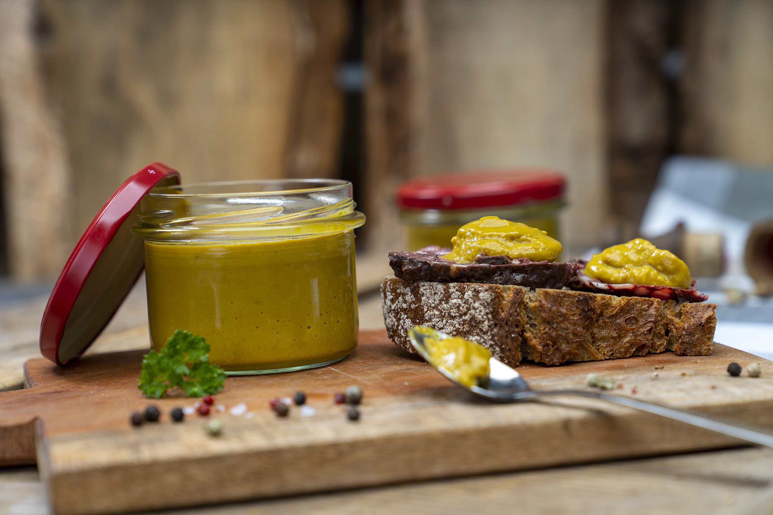 Auf einem Holzbrett ein Glas Senf bzw. Meisters Mostrich. Daneben eine Schnitte mit Rotwurst und Senf. Darum Gewürze und Kräuter. Vorn ein Löffel mit etwas Senf. Hinten ein weiteres Glas.
