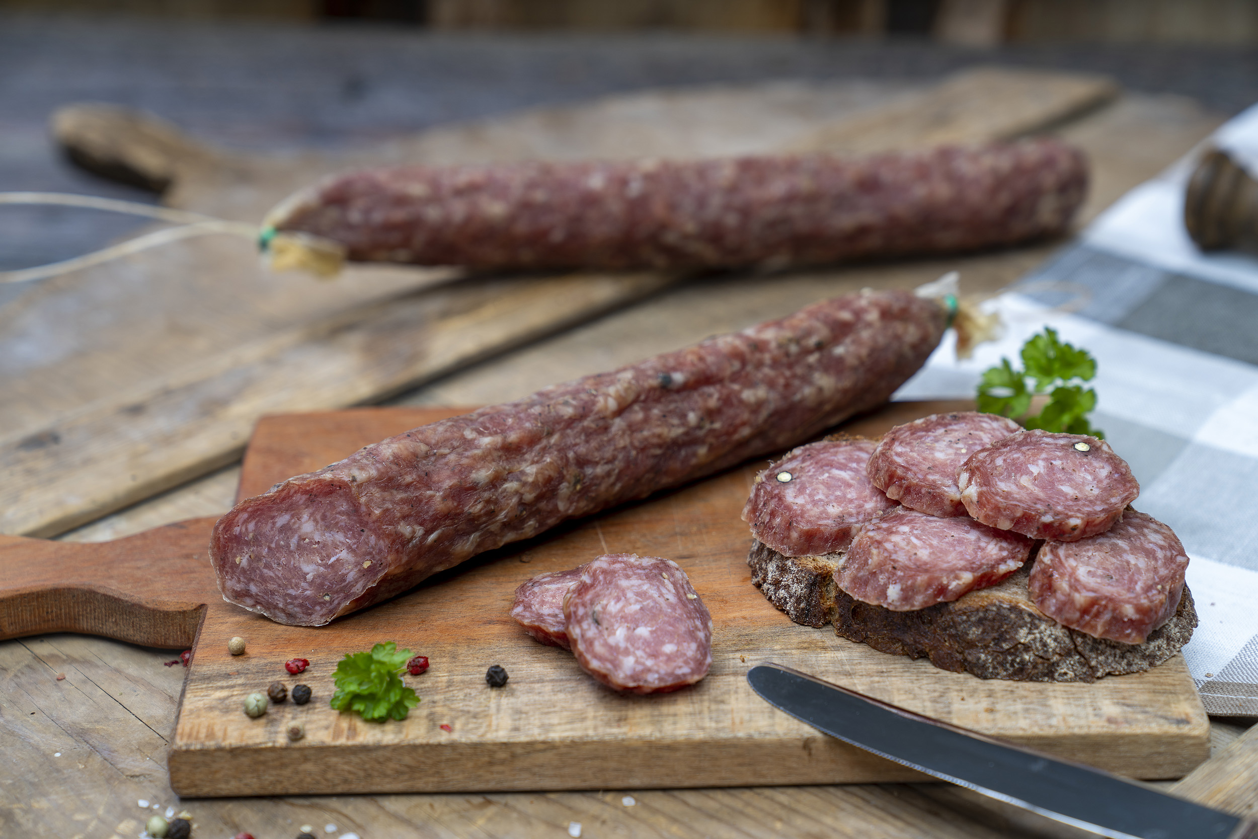 Stracke auf einem Brett. Daneben eine Scheibe Brot mit der Eichsfelder Stracke und Kräuter. Im Hintergrund eine andere Wurst.
