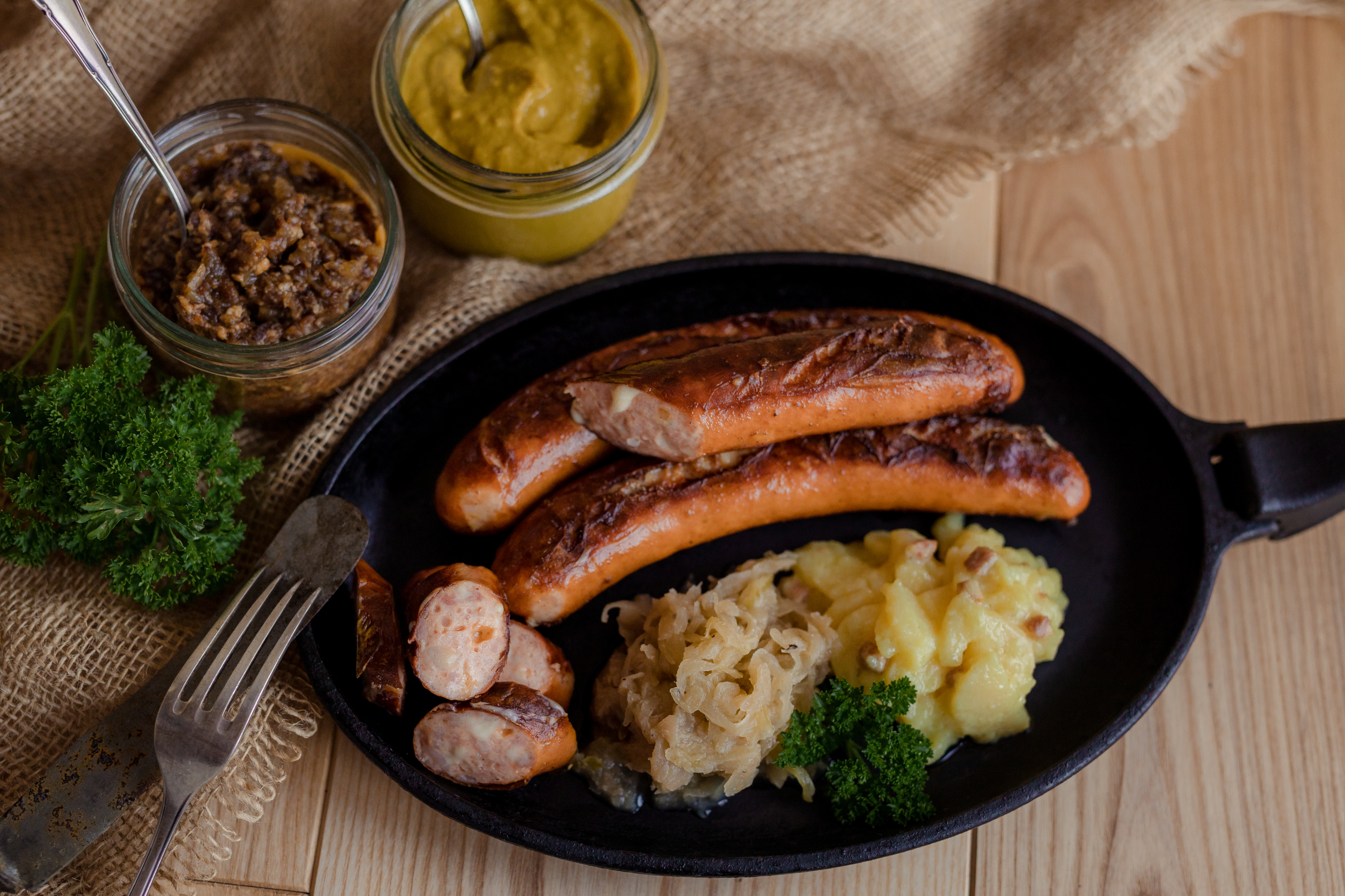 Gebratene Käsekrainer in der Pfanne. Aufgeschnittene Stücke, Sauerkraut und Salatbeilage. Neben der Pfanne Gläser mit Saucen und Kräuter auf Untergrund aus Holz.