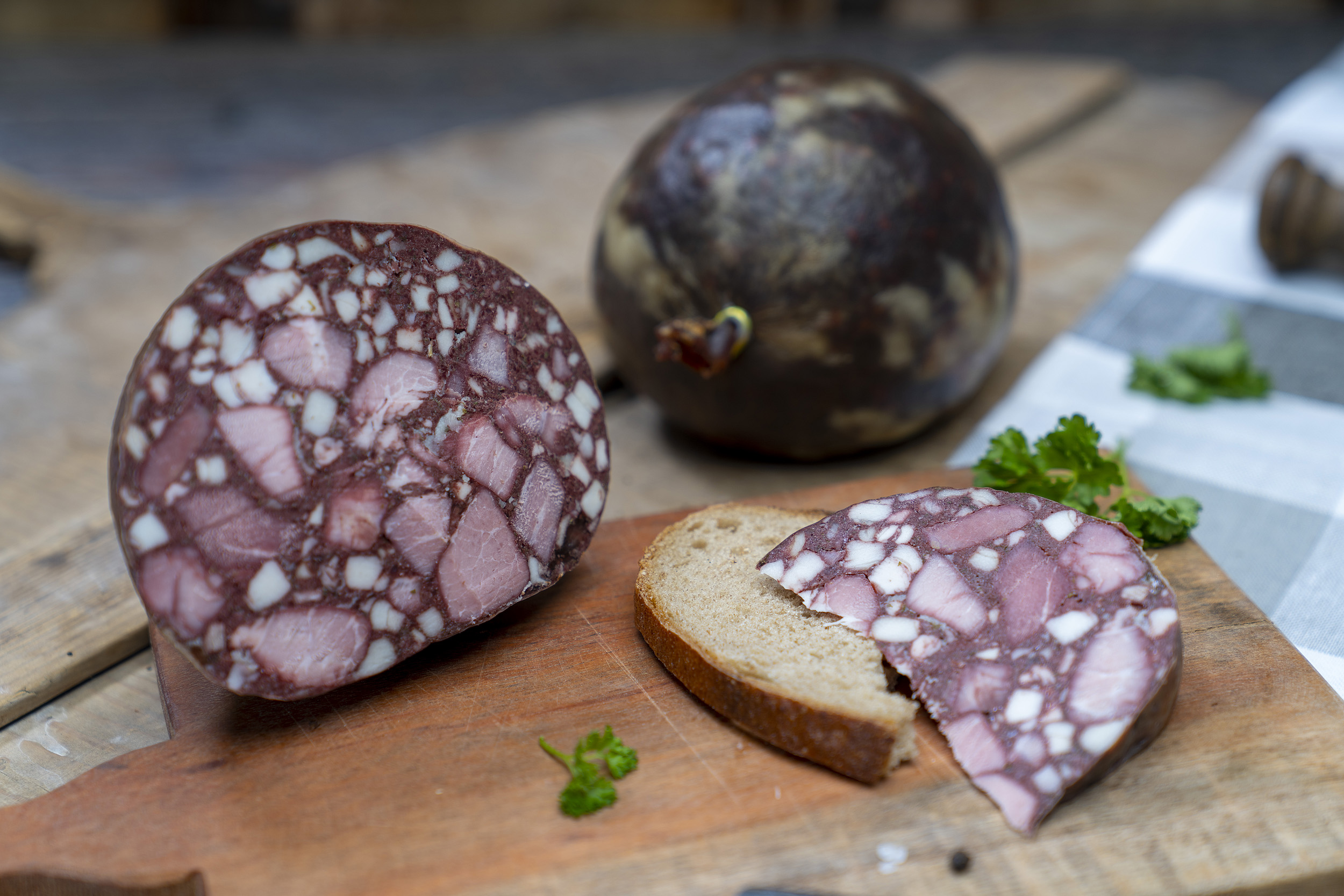 Auf einem Brett eine aufgeschnittene Gutsfleischwurst. Daneben eine Scheibe Brot mit der Wurst. Hinten eine weiters Blase der Wurst.