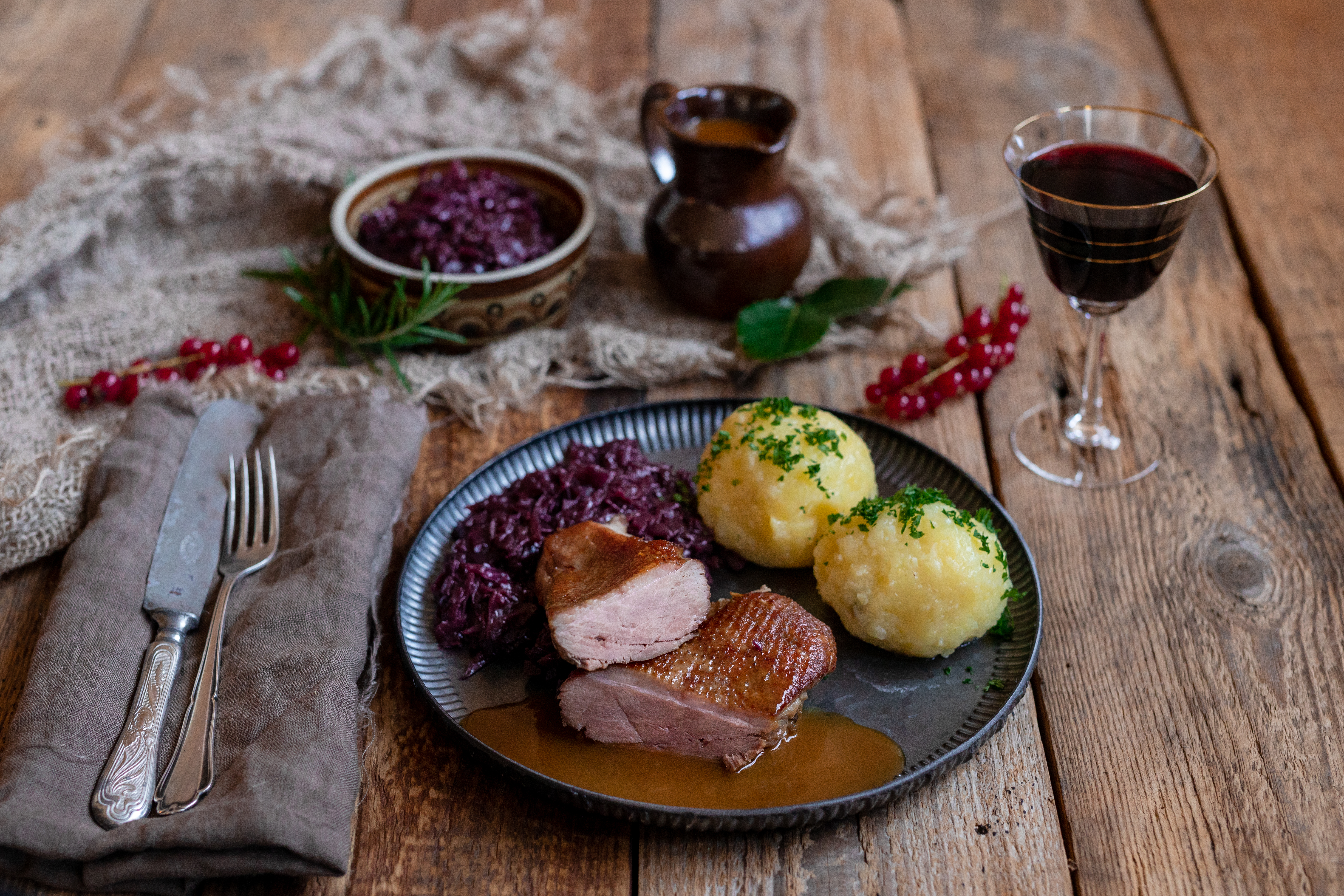 Gänsebrust, Rotkohl und Klöße auf einem Tisch. Daneben Besteck, ein Glas Rotwein und Deko.