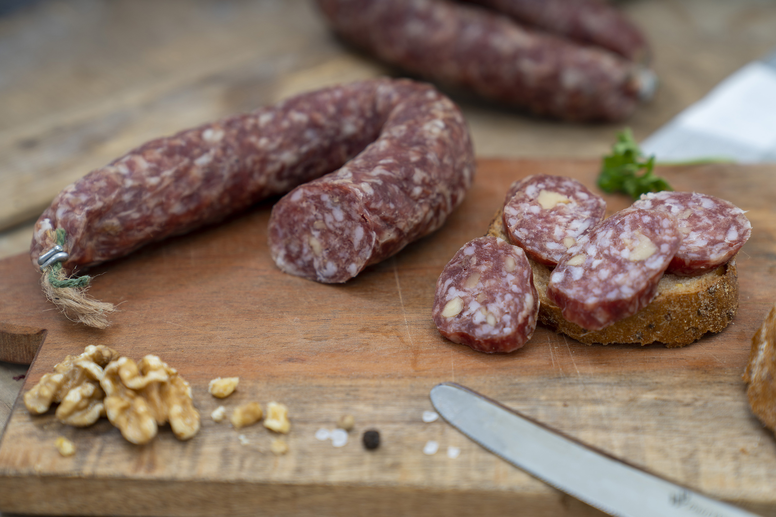 Auf einem Brett eine Walnussschinkenwurst. Daneben Walnüsse, Kräuter und eine Scheibe Brot. Hinten eine weitere Wurst.