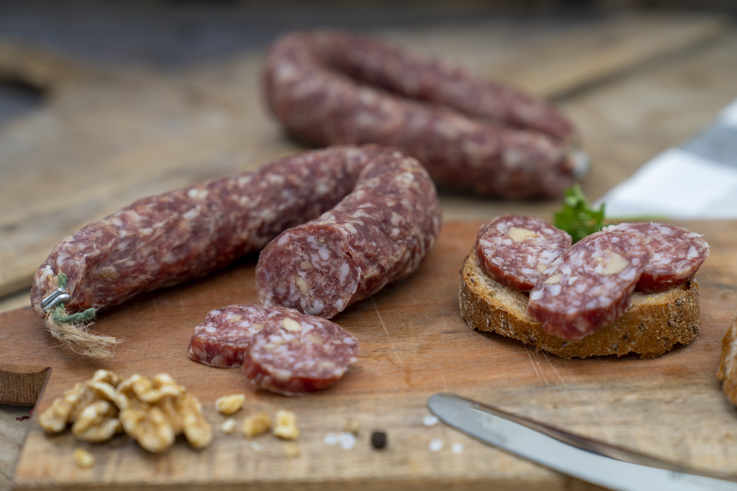 Auf einem Brett eine Walnussschinkenwurst. Daneben Walnüsse, Kräuter und eine Scheibe Brot. Hinten eine weitere Wurst.