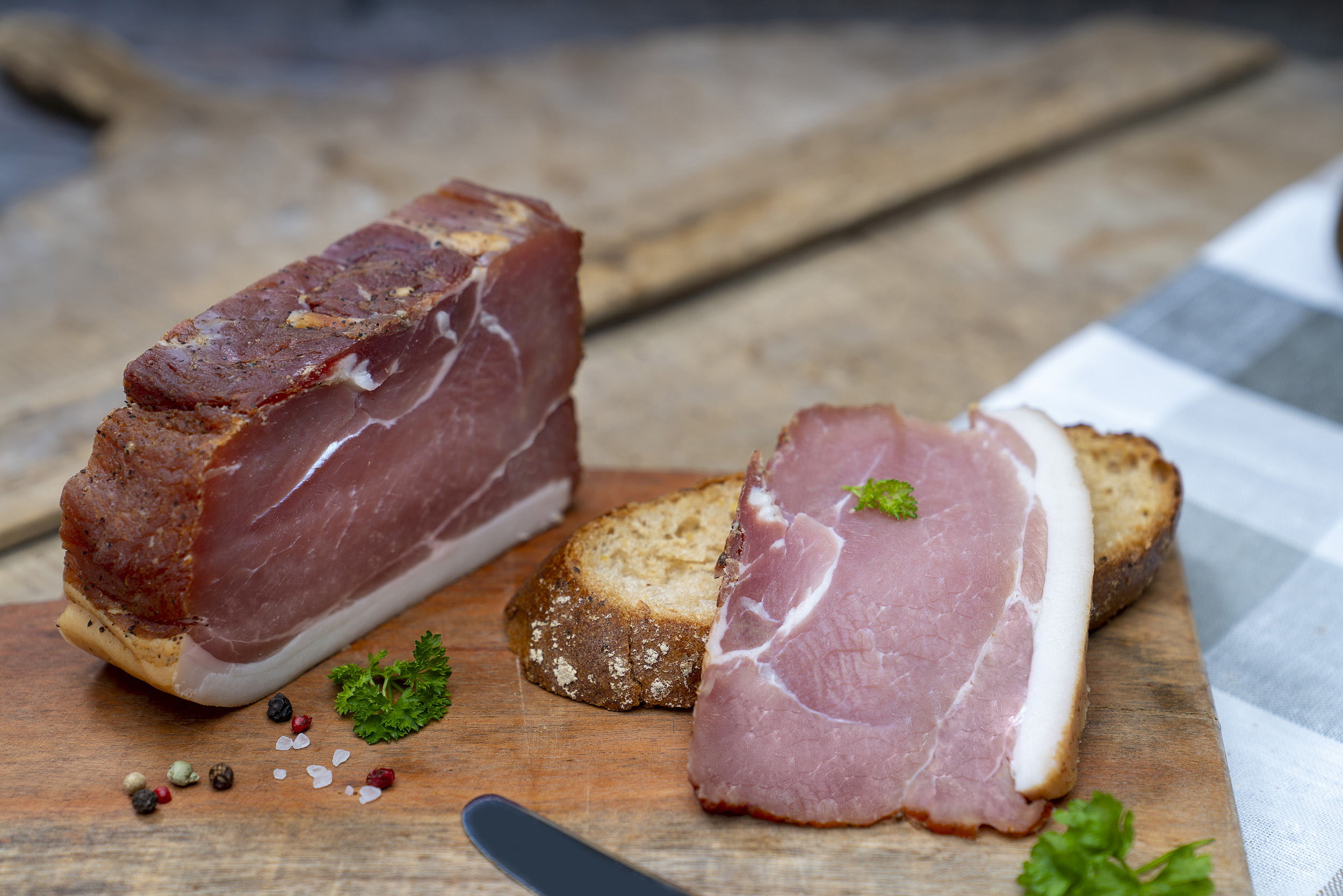 Auf einem Brot eine Scheiben Schinkenspeck. Daneben der Schinken und Gewürze. Im Hintergrund ein weiterer Schinken der Fleischerei Fessel.