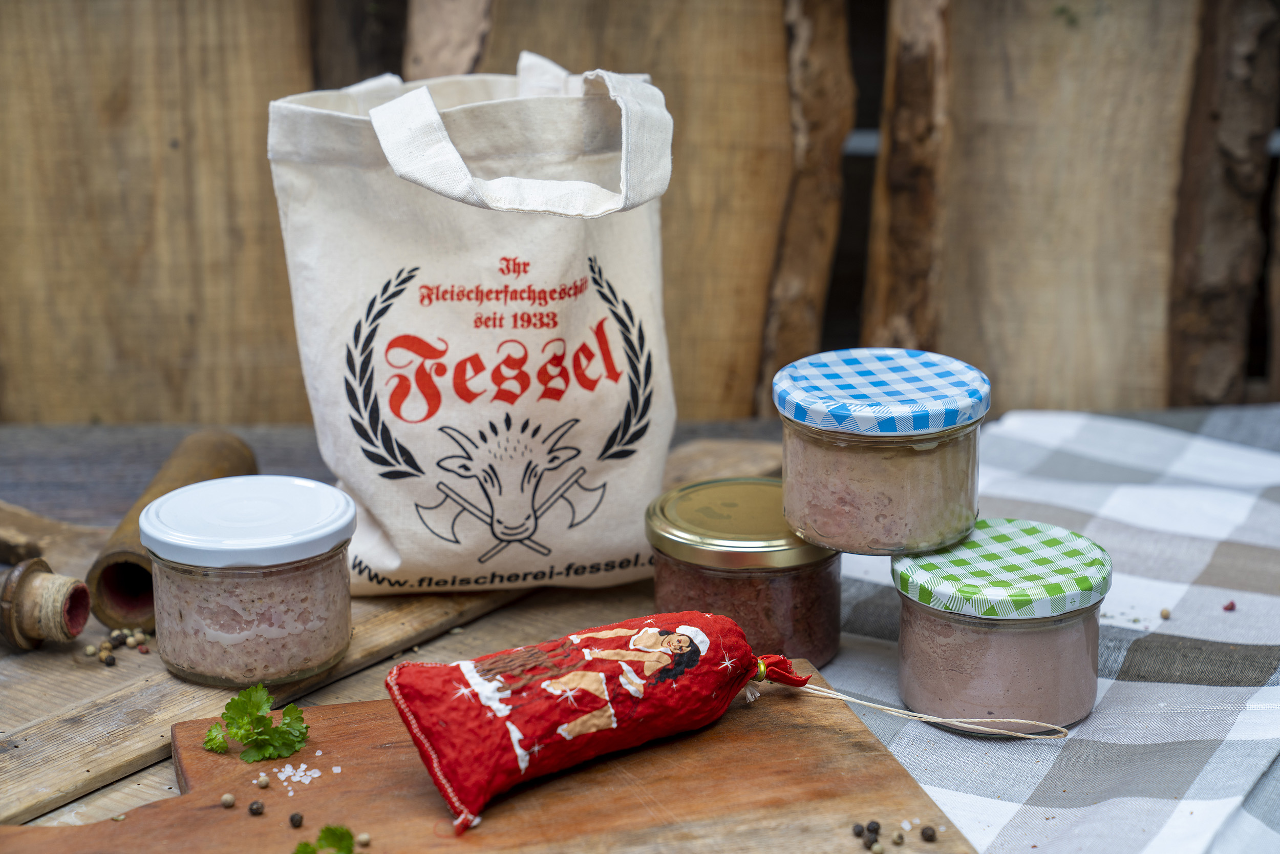 Auf einem Tisch vier verschiedene Konsevengläser Wurstprodukte und eine Salami in weihnachtlichem Look. Hinten ein Jutebeutel der Fleischerei Fessel.