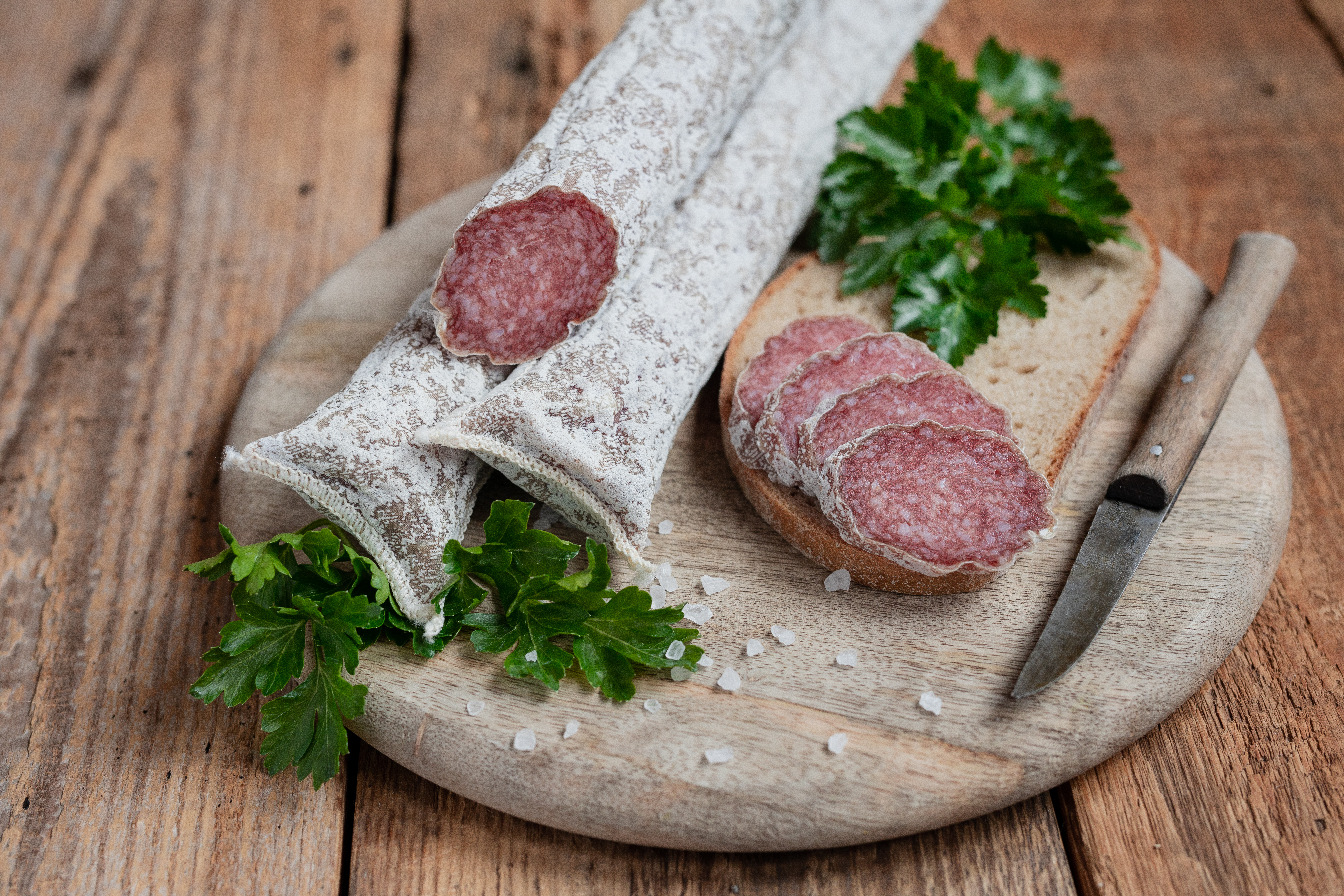 Auf einem Brett zwei mediterrane Salami. Daneben drei Scheiben der Salami, Kräuter und ein Messer.