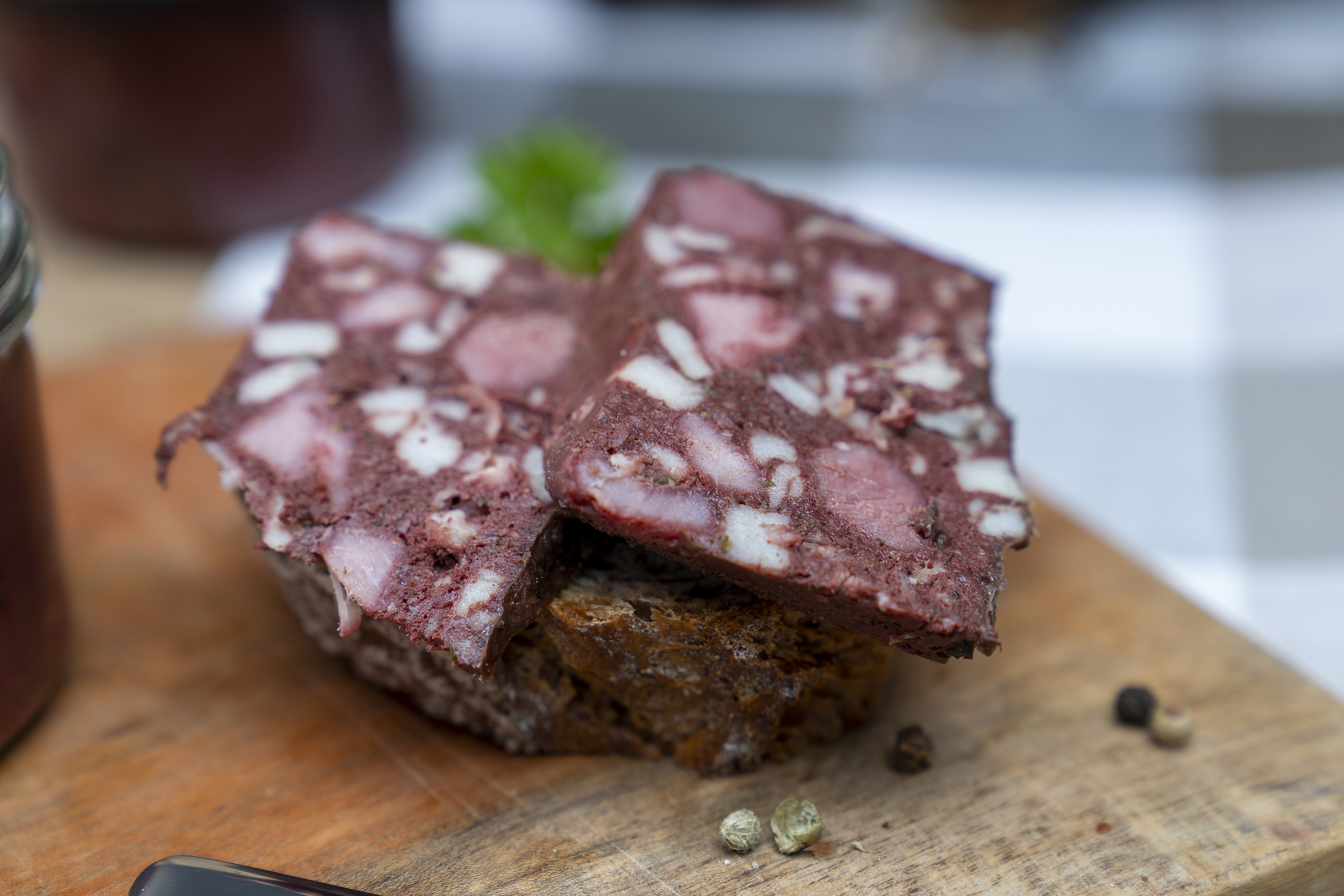 Auf einem Holzbrett steht ein Glas Rotwurst. Daneben eine Scheibe Brot mit Rotwurst Scheiben. Darum Gewürze. Hinten ein weiteres Glas Rotwurst.