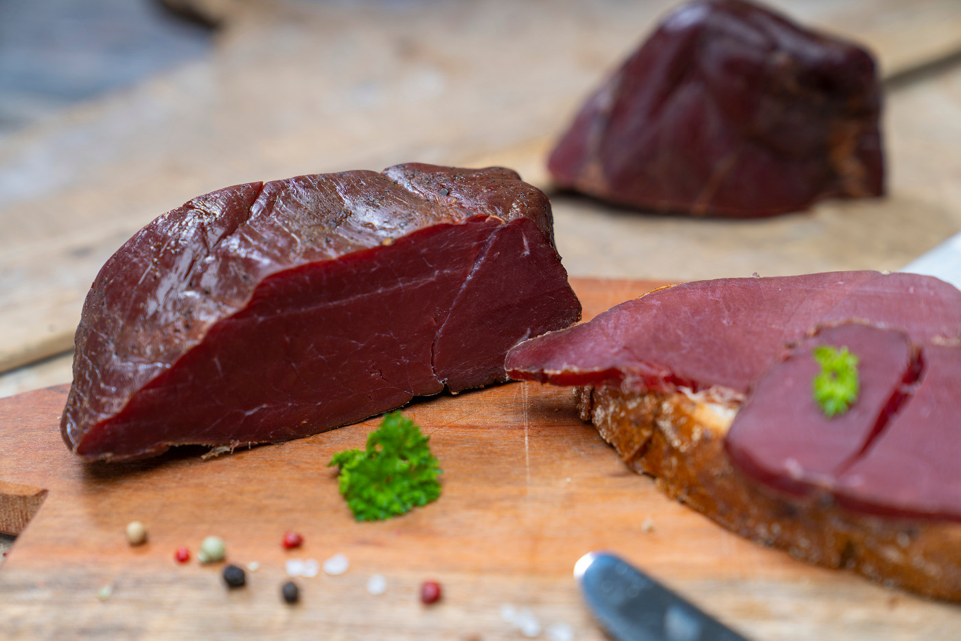 Fessels Wildschinken auf einem Brett. Daneben ein paar Scheiben und Gewürze. Hinten ein weiterer Schinken.