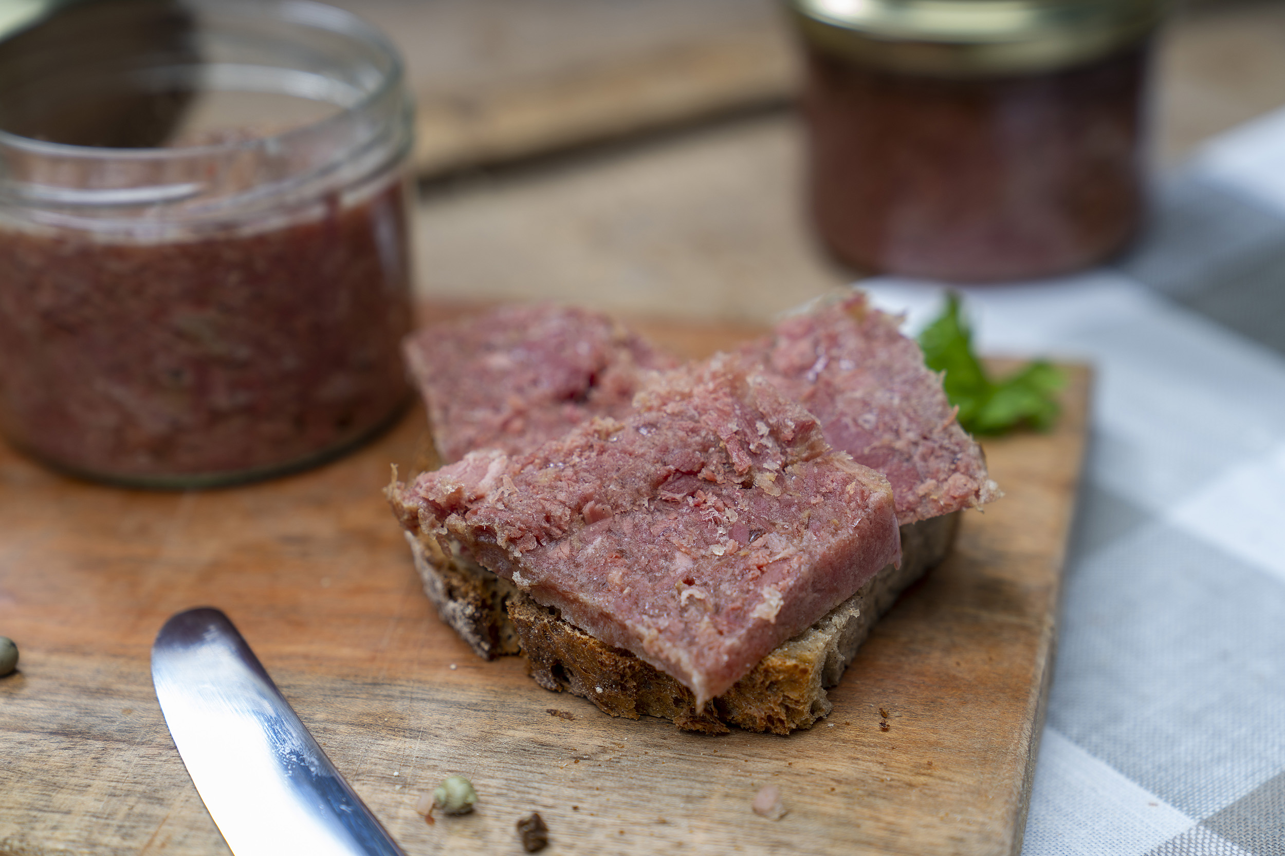 Auf einem Brett ein Glas Corned beef. Daneben ein geschmiertes Brot mit Corned beef. Daneben Gewürze, Kräuter und ein Messer. Im Hintergrund ein weiteres Glas.