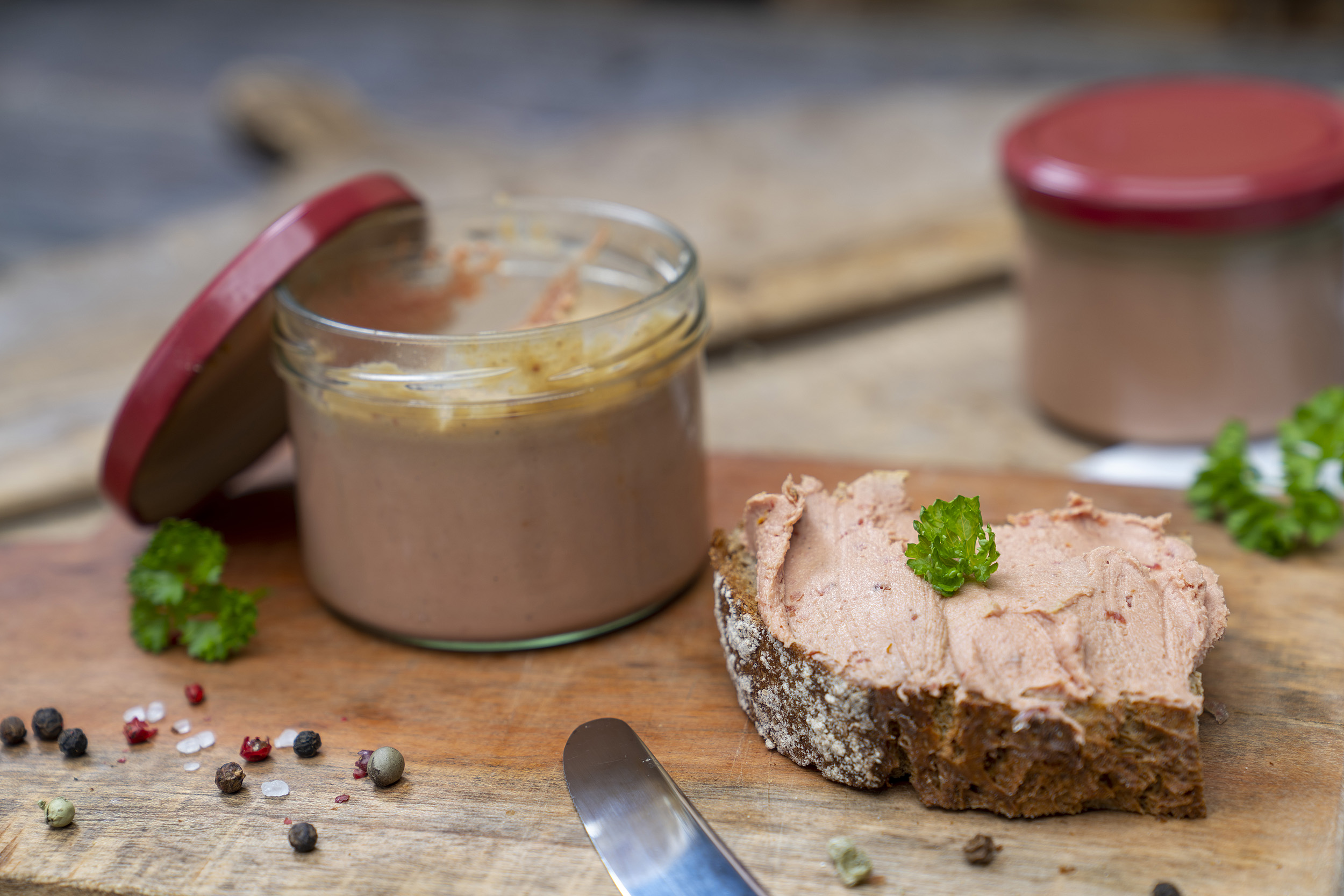 Auf einem Holzbrett ein Glas Tomatenleberwurst. Daneben eine geschmierte Schnitte und Kräuter. Im Hintergrund noch ein Glas.