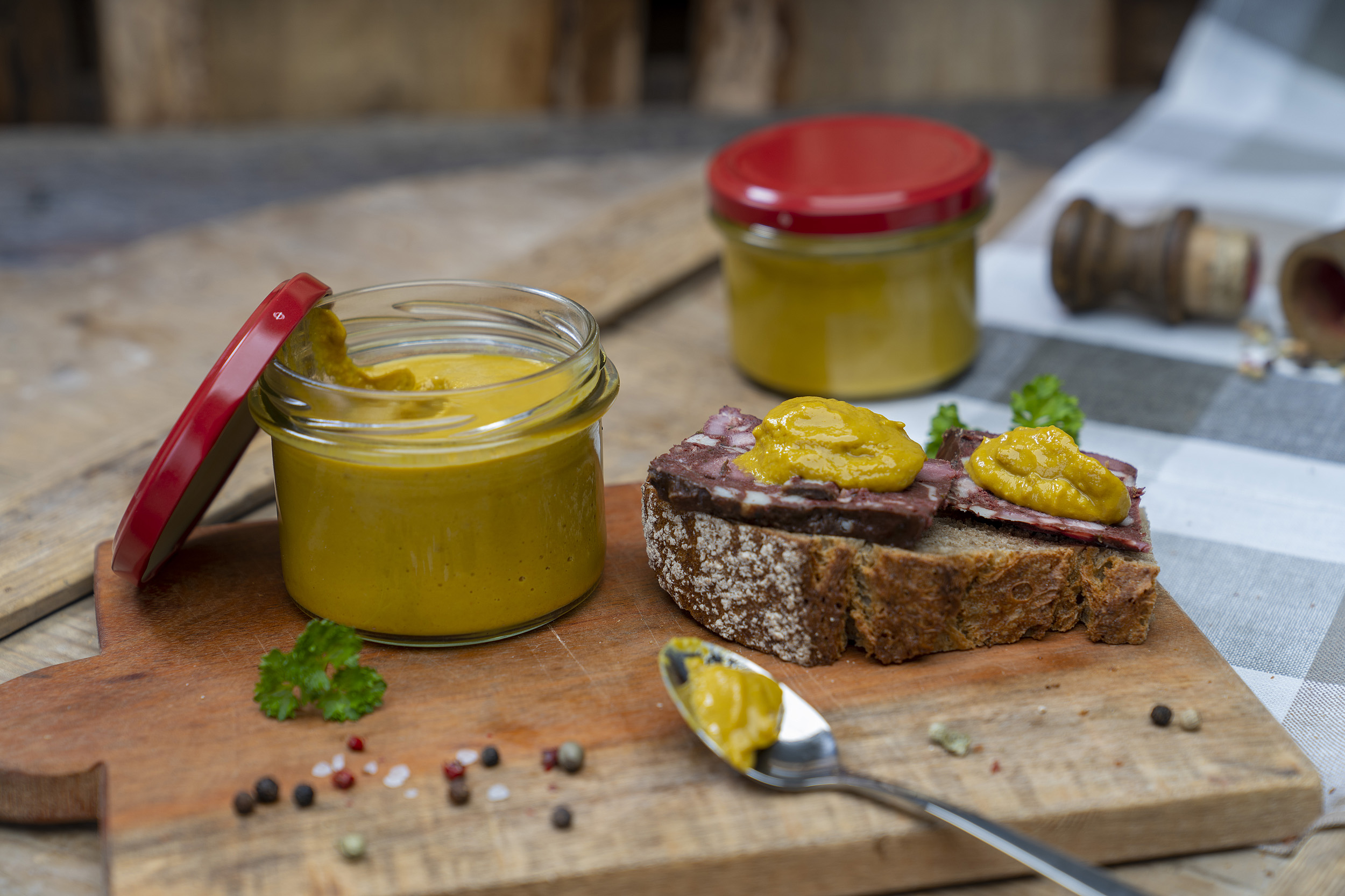 Auf einem Holzbrett ein Glas Senf bzw. Meisters Mostrich. Daneben eine Schnitte mit Rotwurst und Senf. Darum Gewürze und Kräuter. Vorn ein Löffel mit etwas Senf. Hinten ein weiteres Glas.