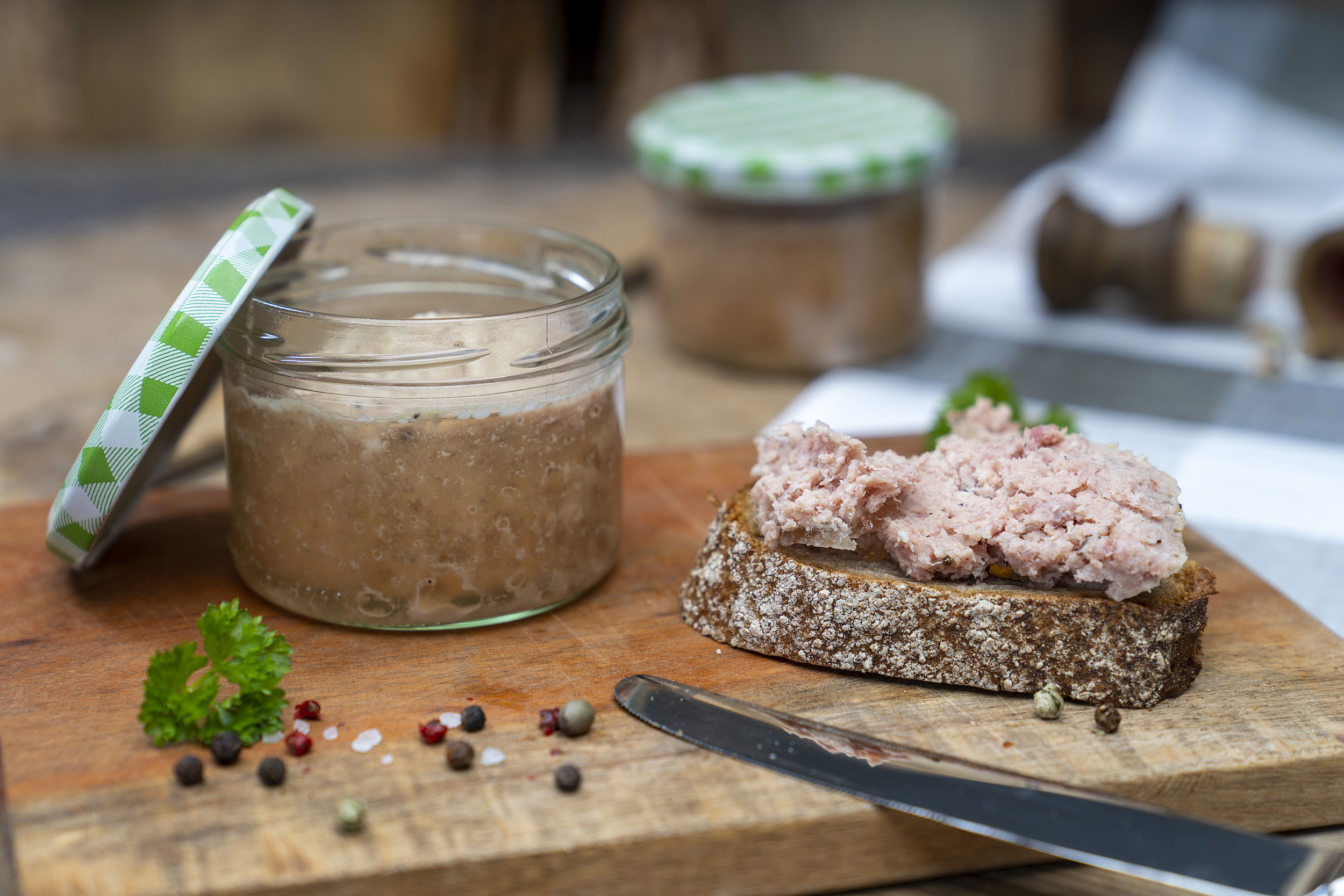 Auf einem Holzbrett steht ein Glas Schartenwurst. Daneben eine Scheibe Brot mit der Wurst. Drum herum Kräuter und Gewürze. Hinten ein weiteres Glas.