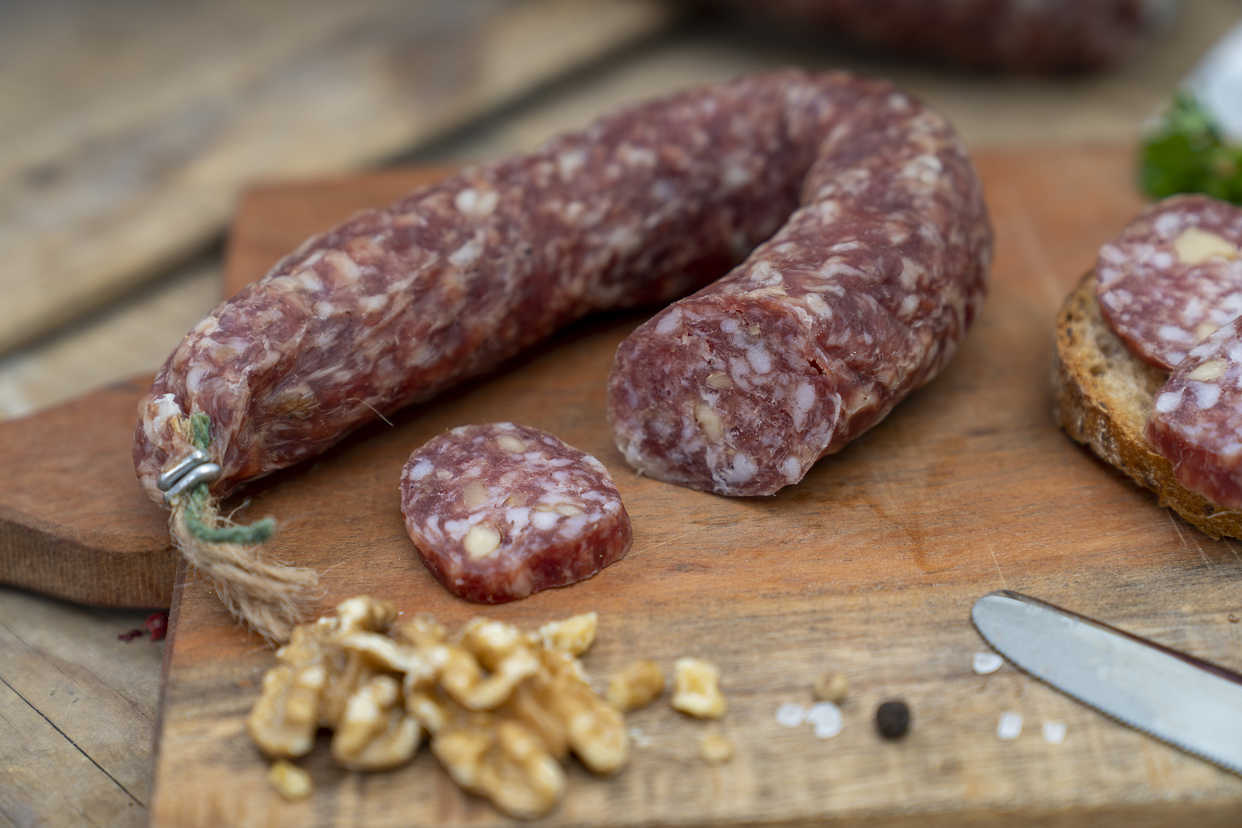 Auf einem Brett eine Walnussschinkenwurst. Daneben Walnüsse, Kräuter und eine Scheibe Brot.