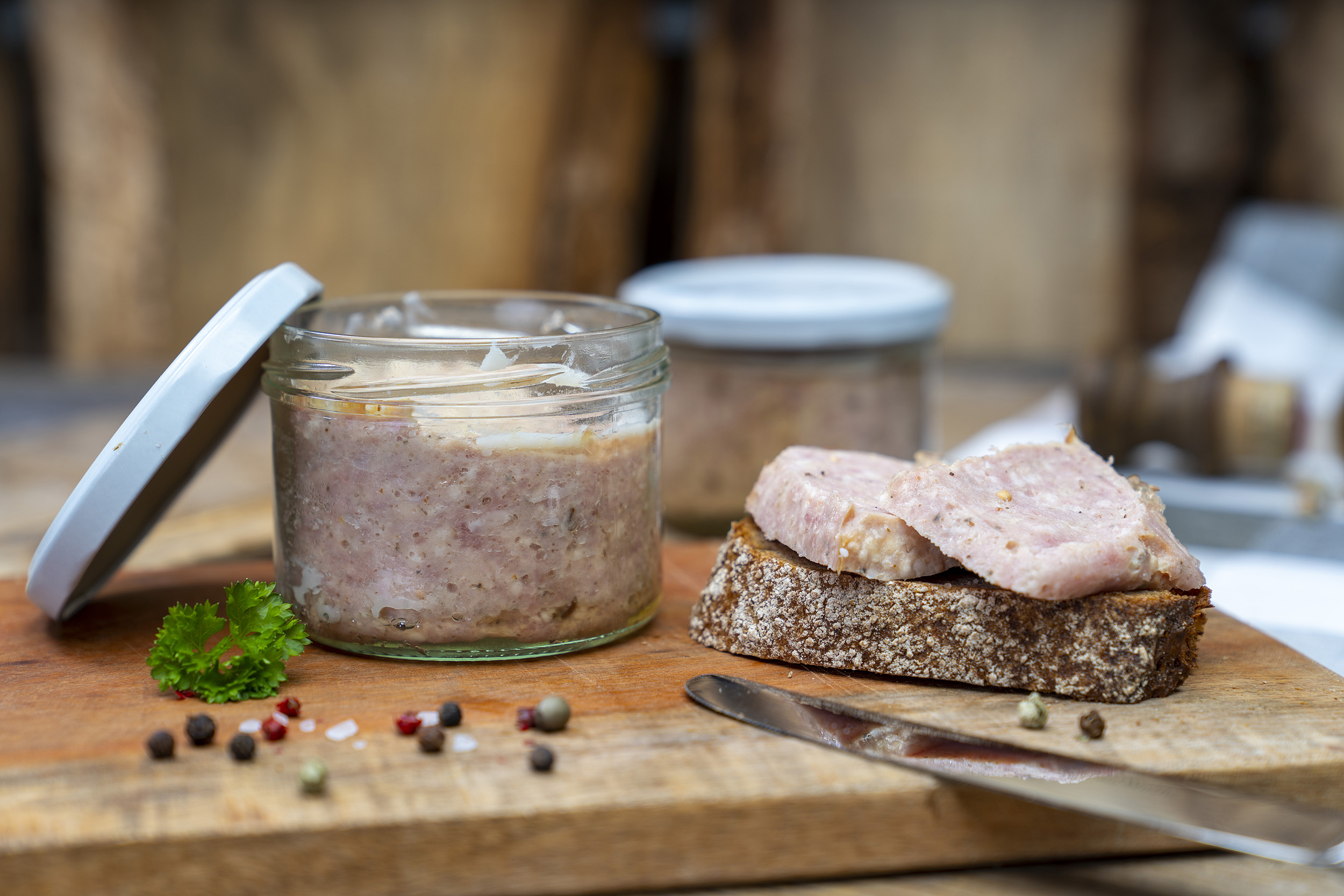 Auf einem Holzbrett ein Glas mit Mettwurst. Daneben eine Schnitte mit Mettwurst, Kräuter und Gewürze. Im Hintergrund steht ein weiteres Glas. Vorn lehnt ein Messer am Brett.